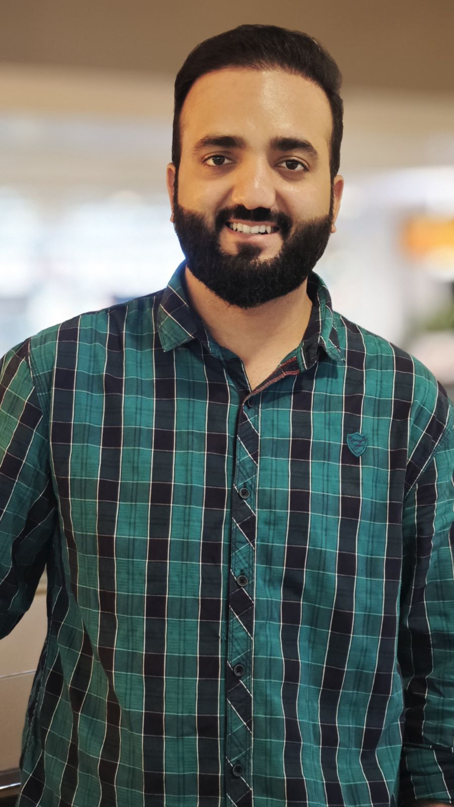 Man with dark hair and beard smiling, wearing a green and blue plaid shirt, standing indoors.
