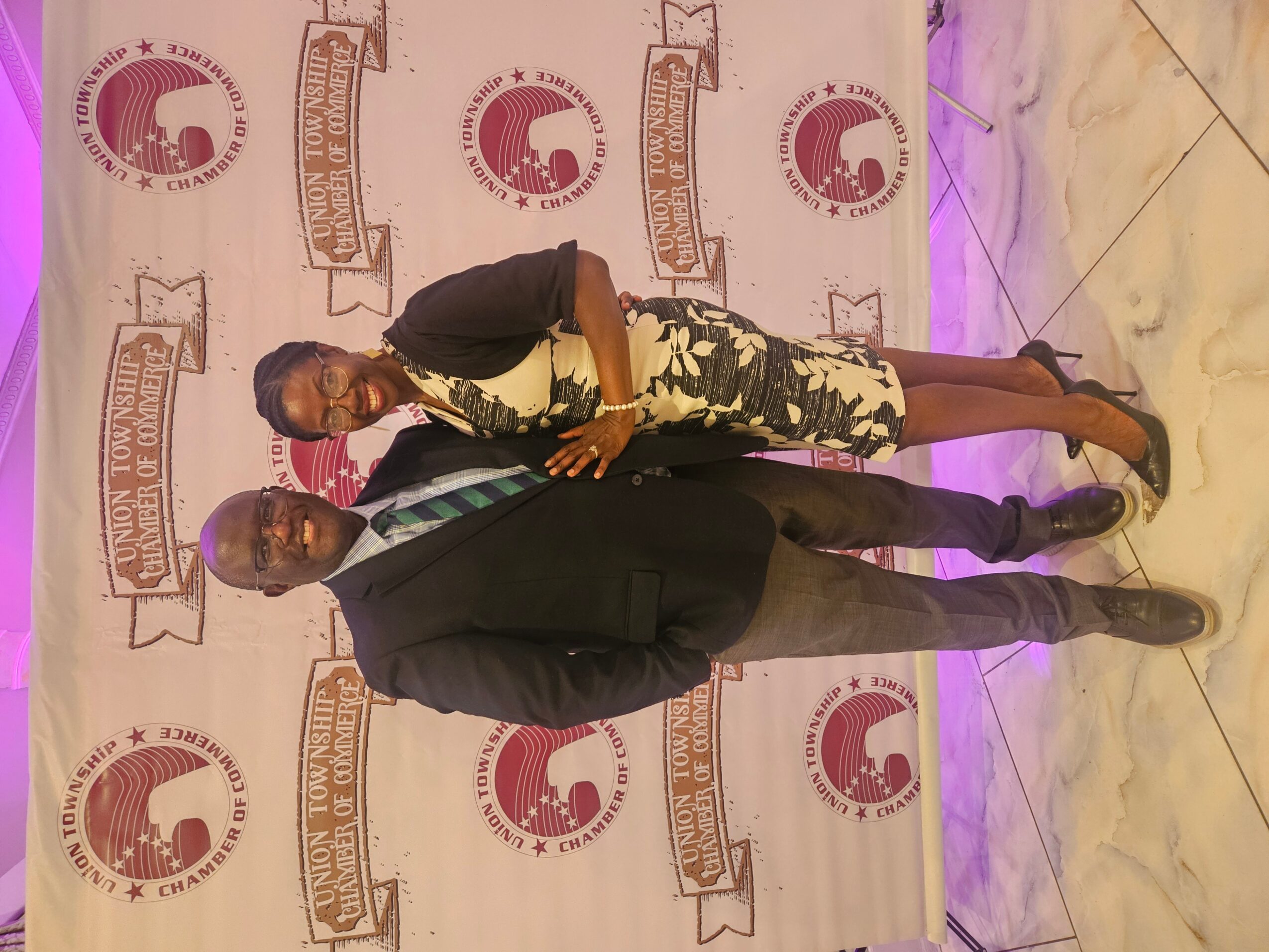 Two people standing in front of a backdrop with logos, one in a suit and the other in a floral dress.