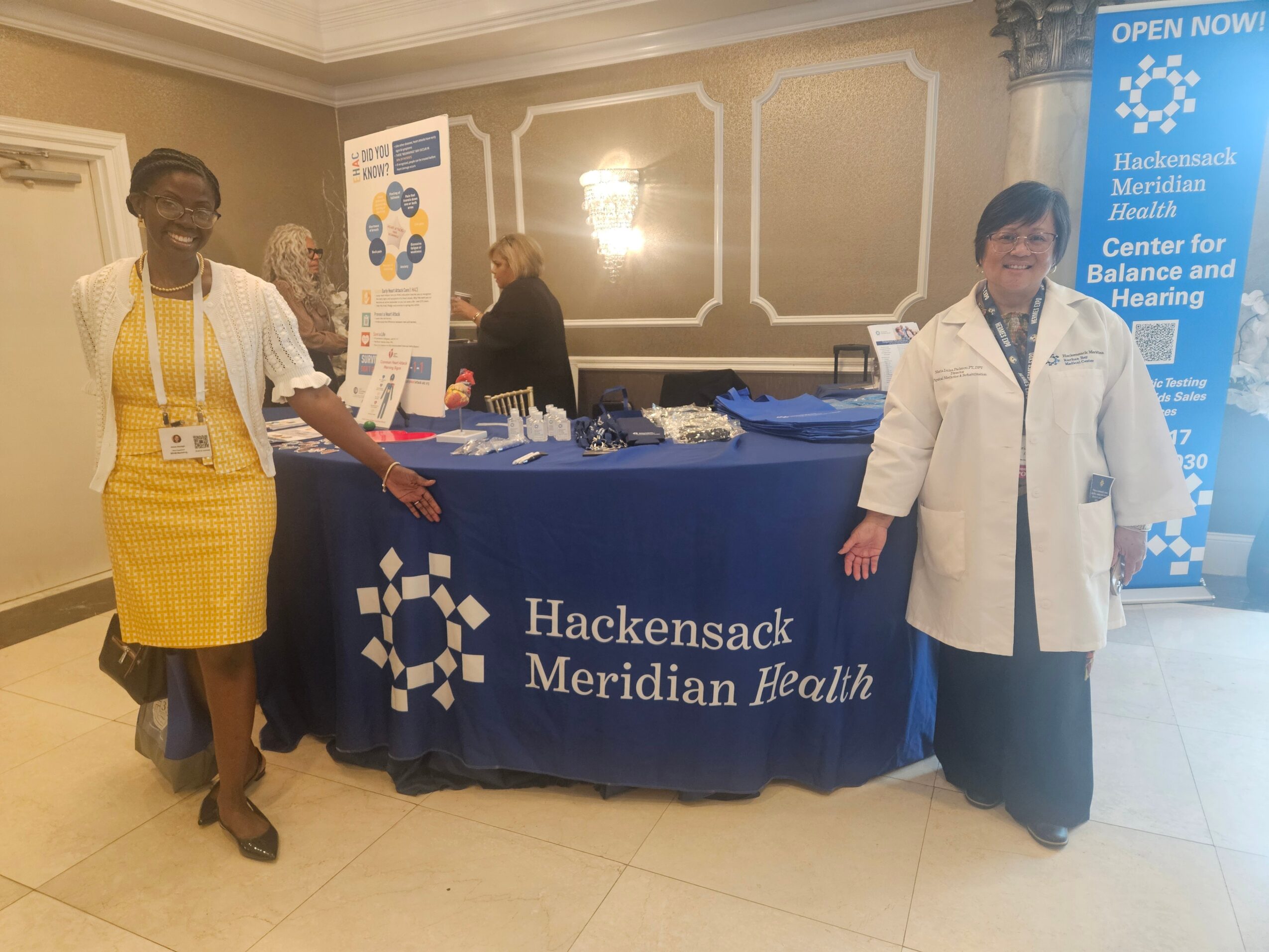 Two women stand beside a table with a Hackensack Meridian Health banner, in a decorated room with beige walls and lighting.