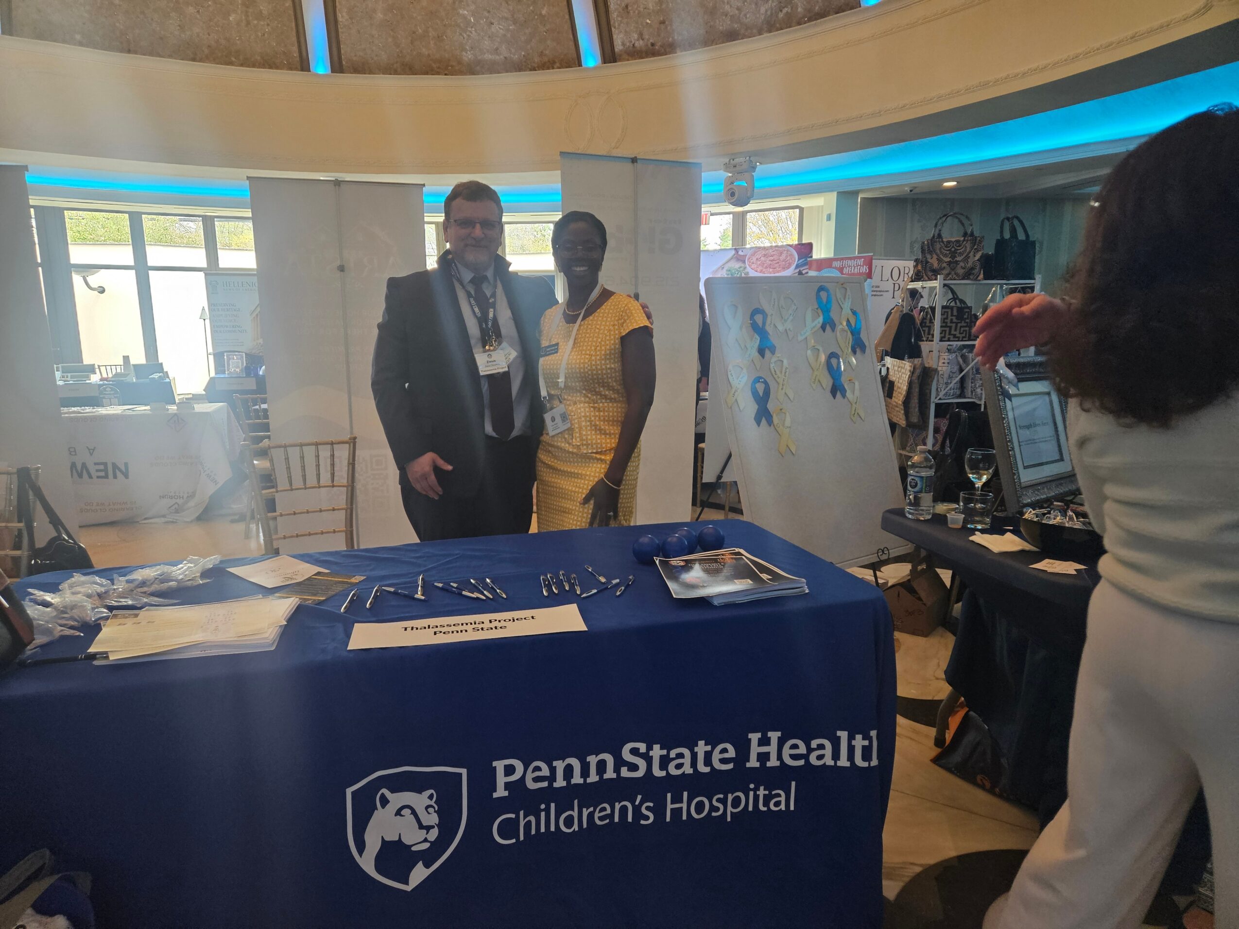 Two people standing behind a table with Penn State Health Children's Hospital logo, in a bright indoor setting.