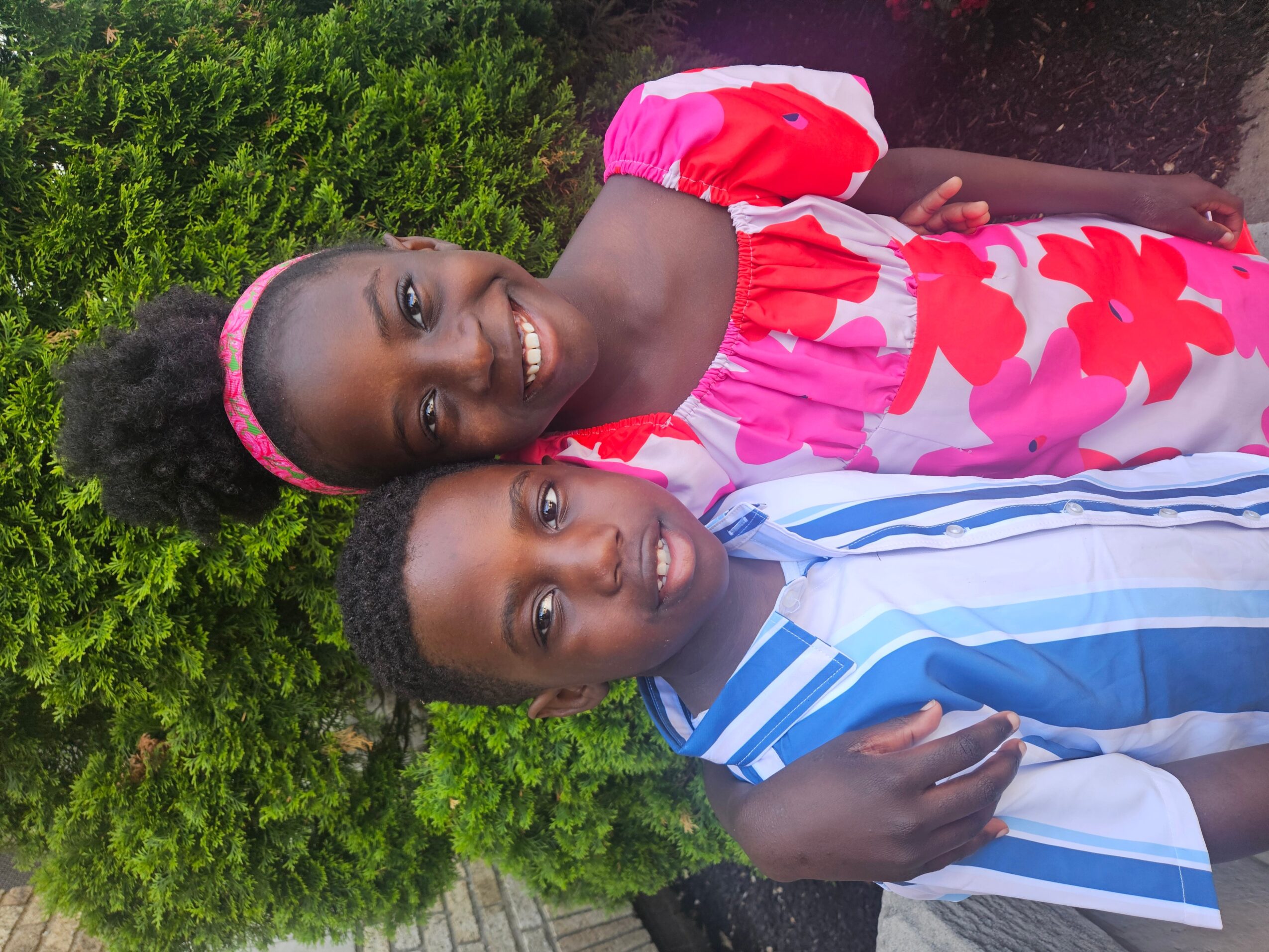 Two children smiling outdoors, girl in pink and white dress, boy in blue and white striped shirt, green bushes in background.