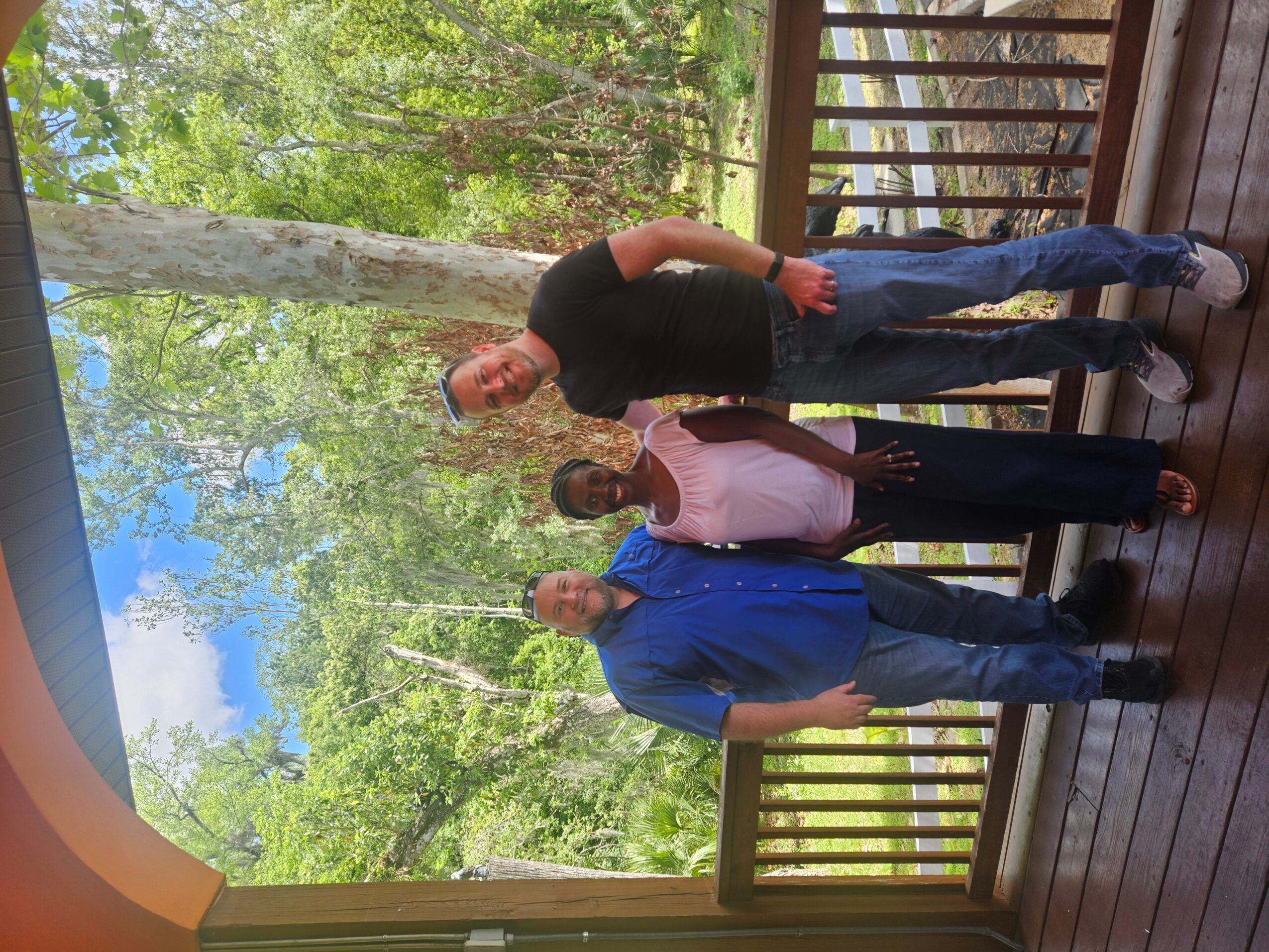 Three people standing on a wooden deck with trees and blue sky in the background.