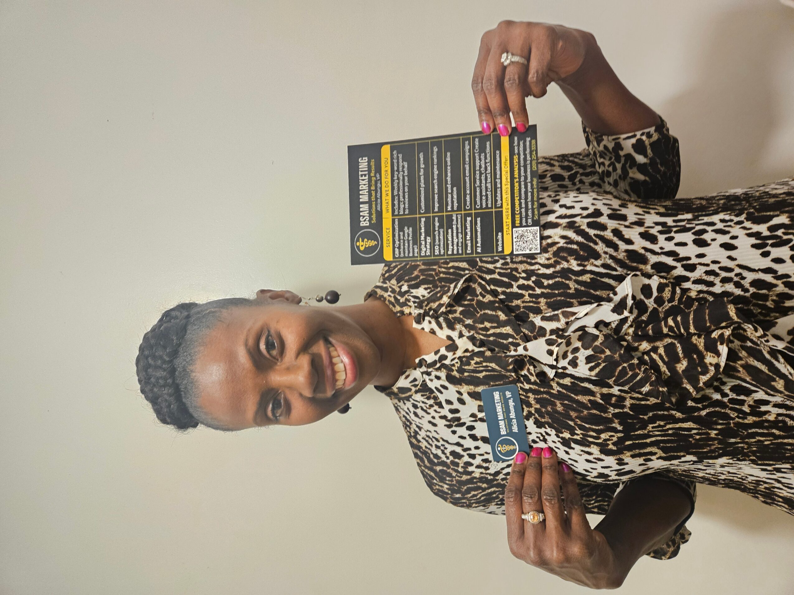 Woman with short hair holding a product box, smiling, wearing a leopard print top, standing against a plain wall.
