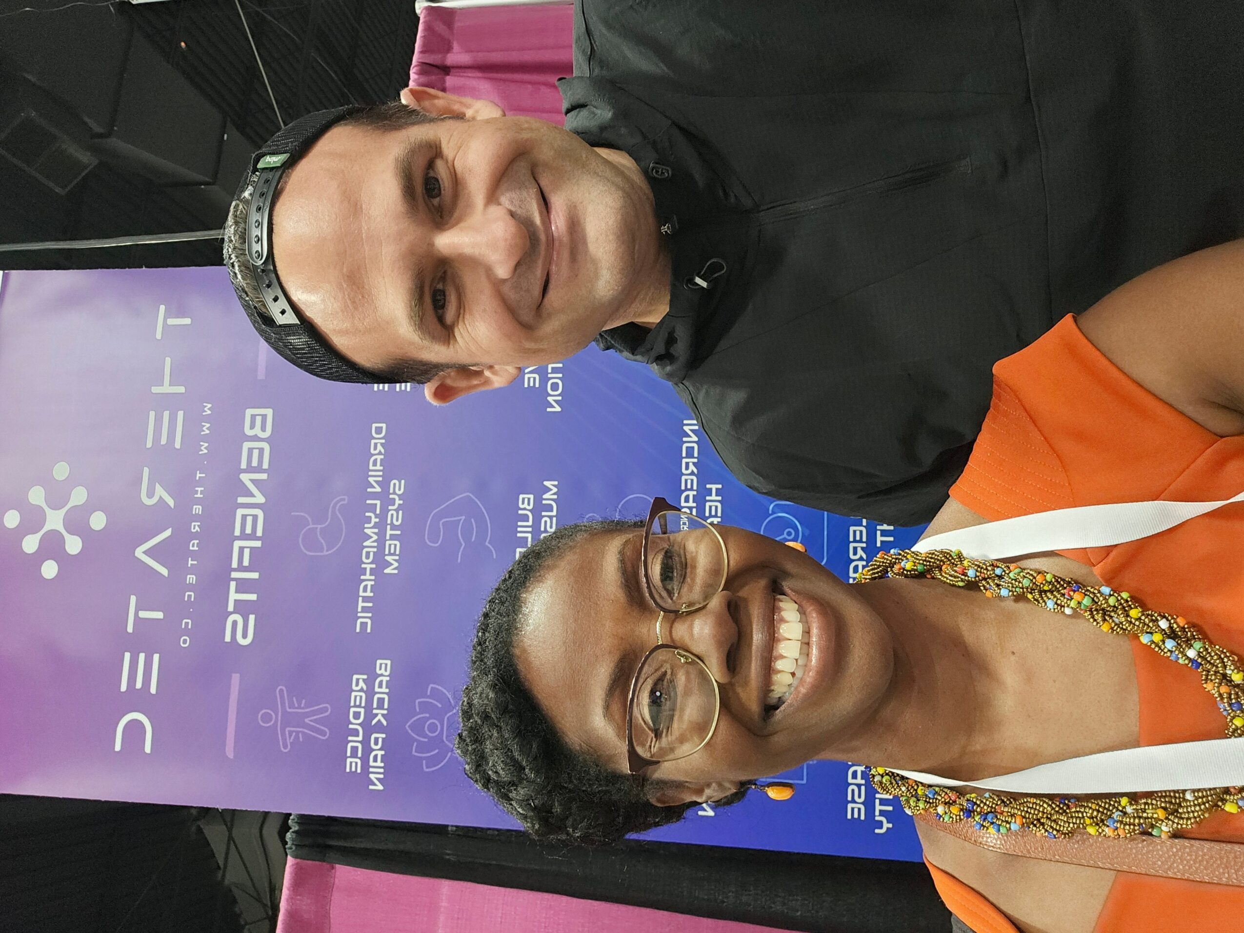 Two people smiling, one wearing glasses and a colorful necklace, the other in a black shirt, standing in front of a purple and blue background.