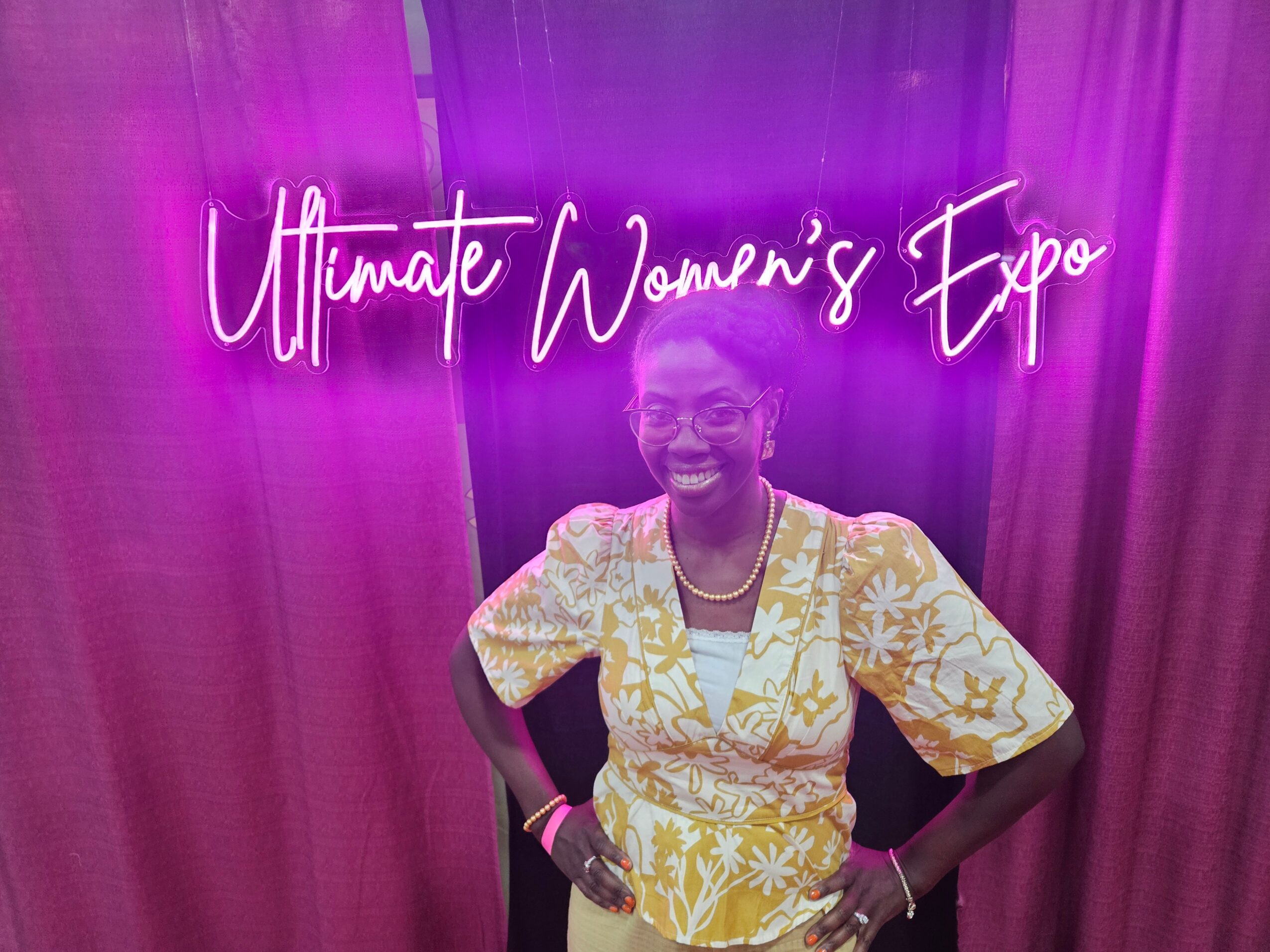 Smiling woman in yellow and white dress standing in front of a neon sign that reads 'Ultimate Women's Expo'.