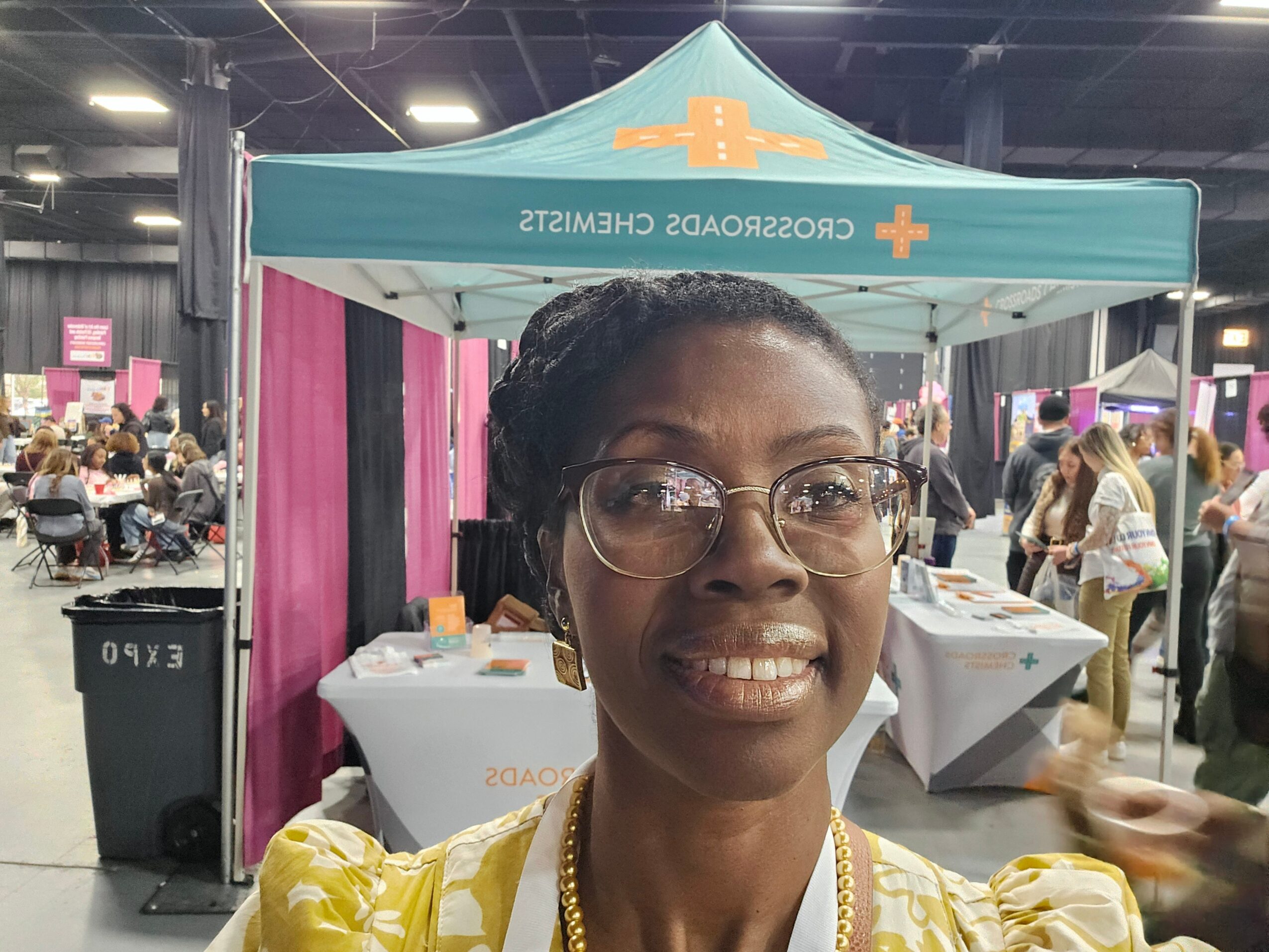 Woman with glasses smiling in front of a health booth at an indoor event, with tables and people in the background.