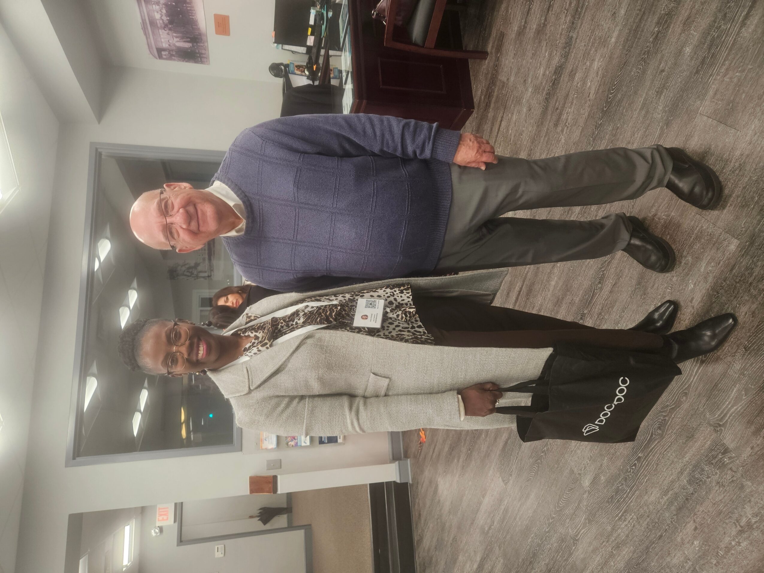 Two people standing indoors, one in a blue sweater and gray pants, the other in a white coat and black shoes.