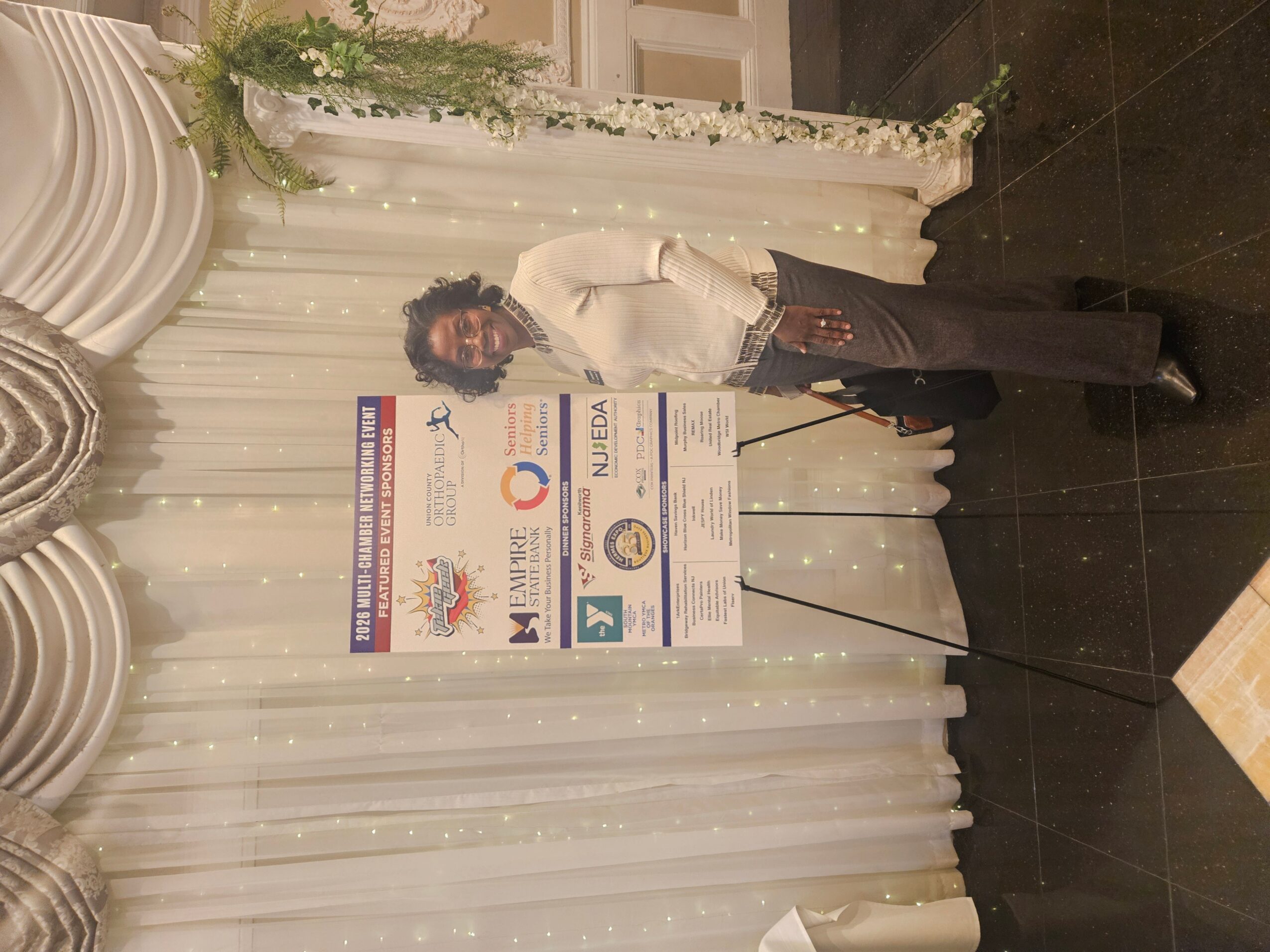 Person standing next to a large poster board at an event, with white drapes and floral decorations in the background.