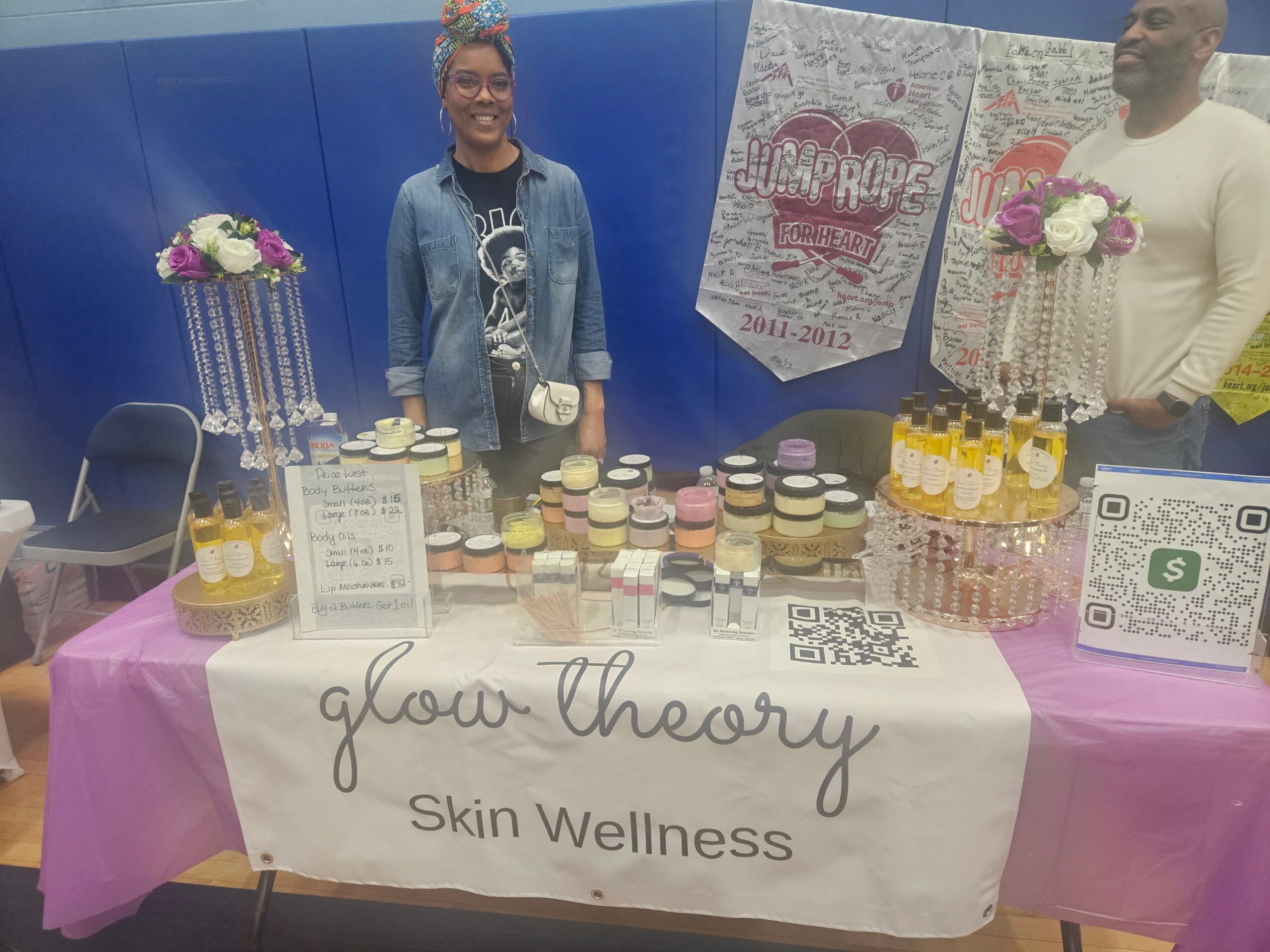 Two people stand behind a table with cupcakes and floral decorations, promoting Glow Theory Skin Wellness, with a pink tablecloth and a banner.