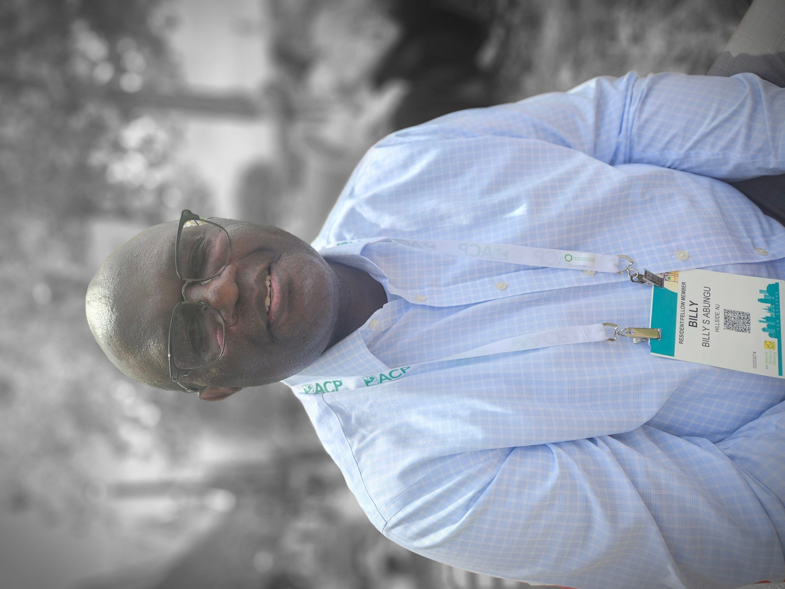 Man wearing glasses and a light blue shirt outdoors, smiling, with trees in the background.