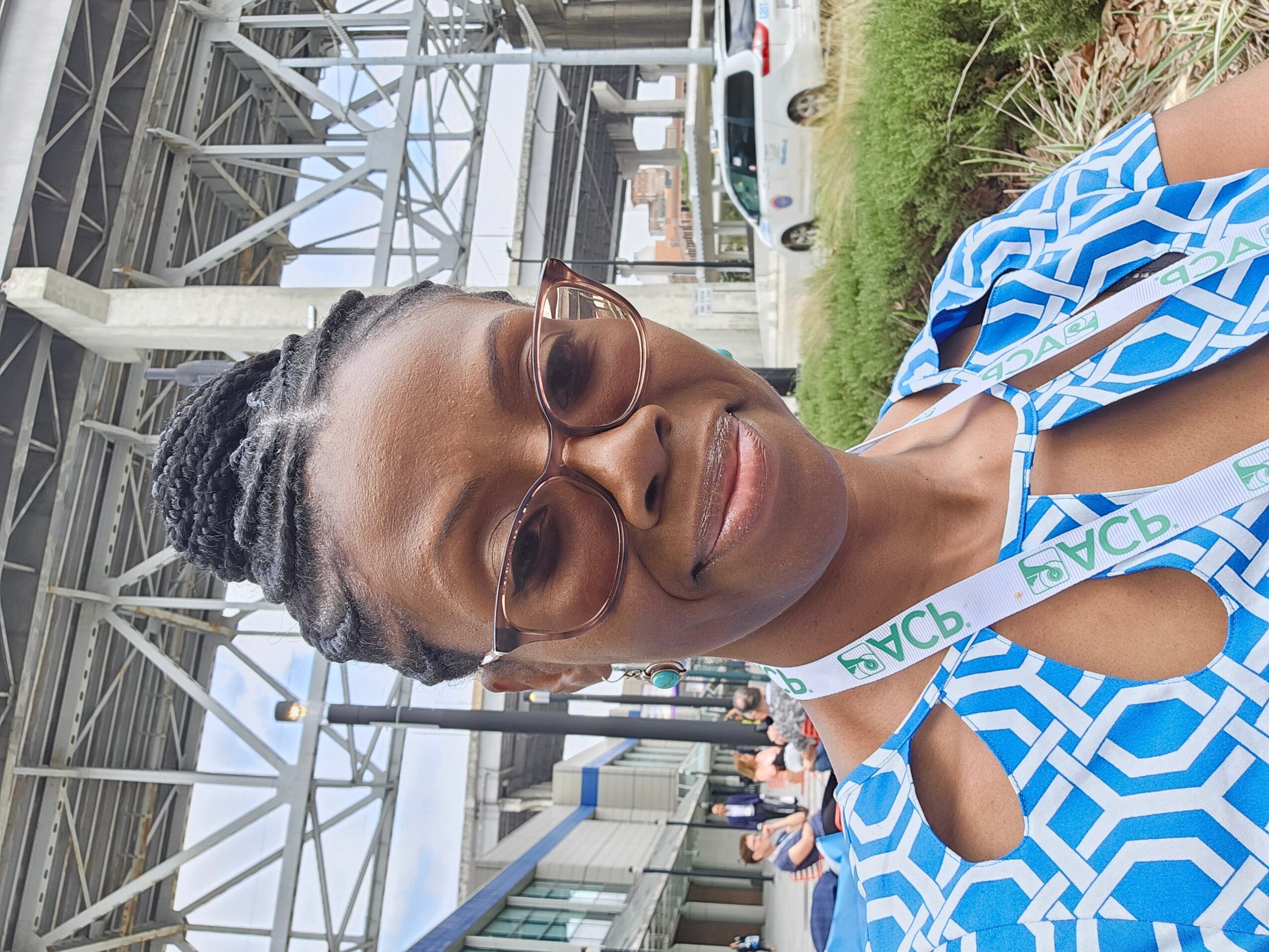 Woman with sunglasses smiling outdoors, wearing a blue and white patterned dress, under a metal structure.