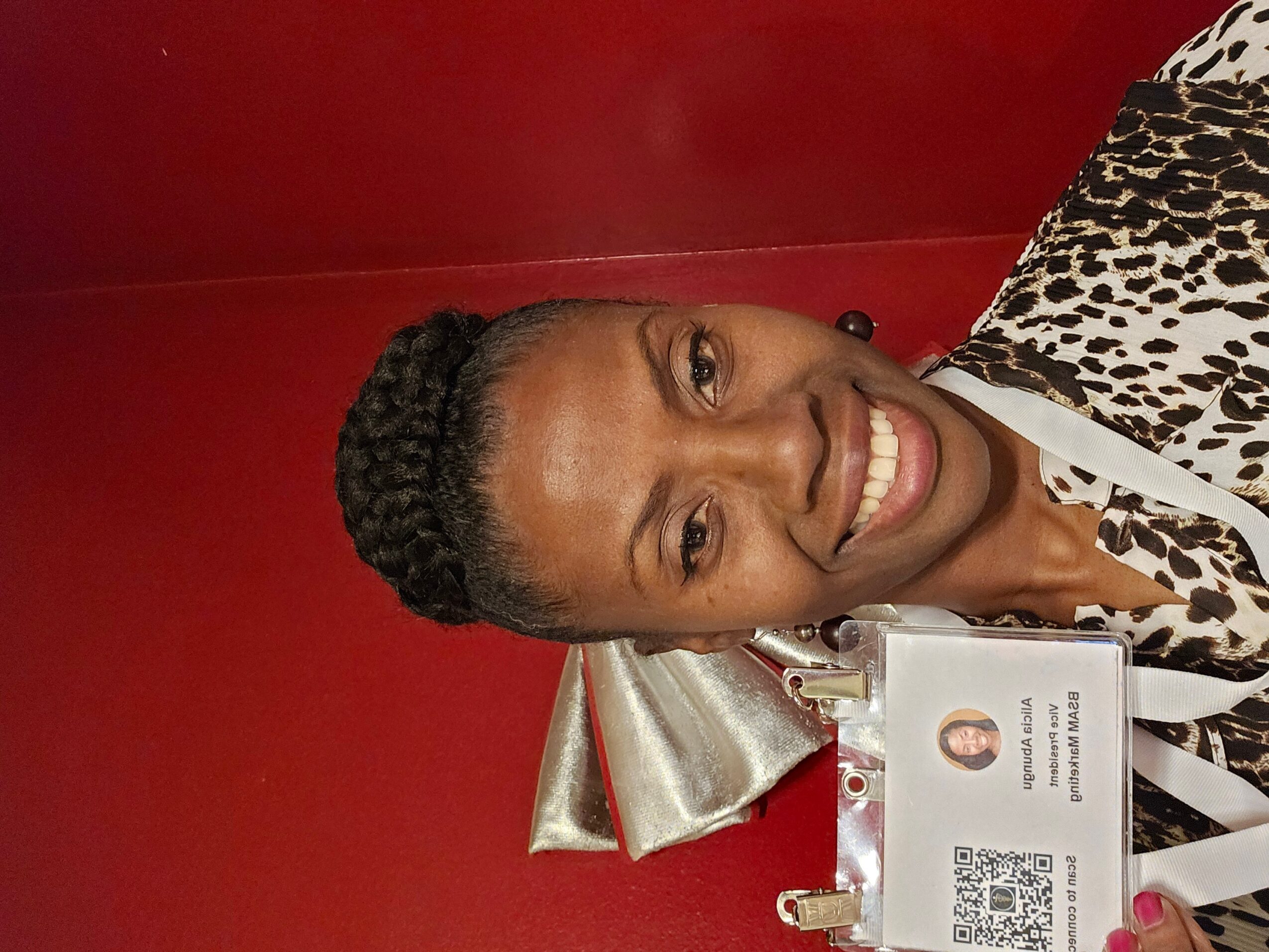 Woman smiling, wearing a patterned top, with a red background and metallic decoration behind her.