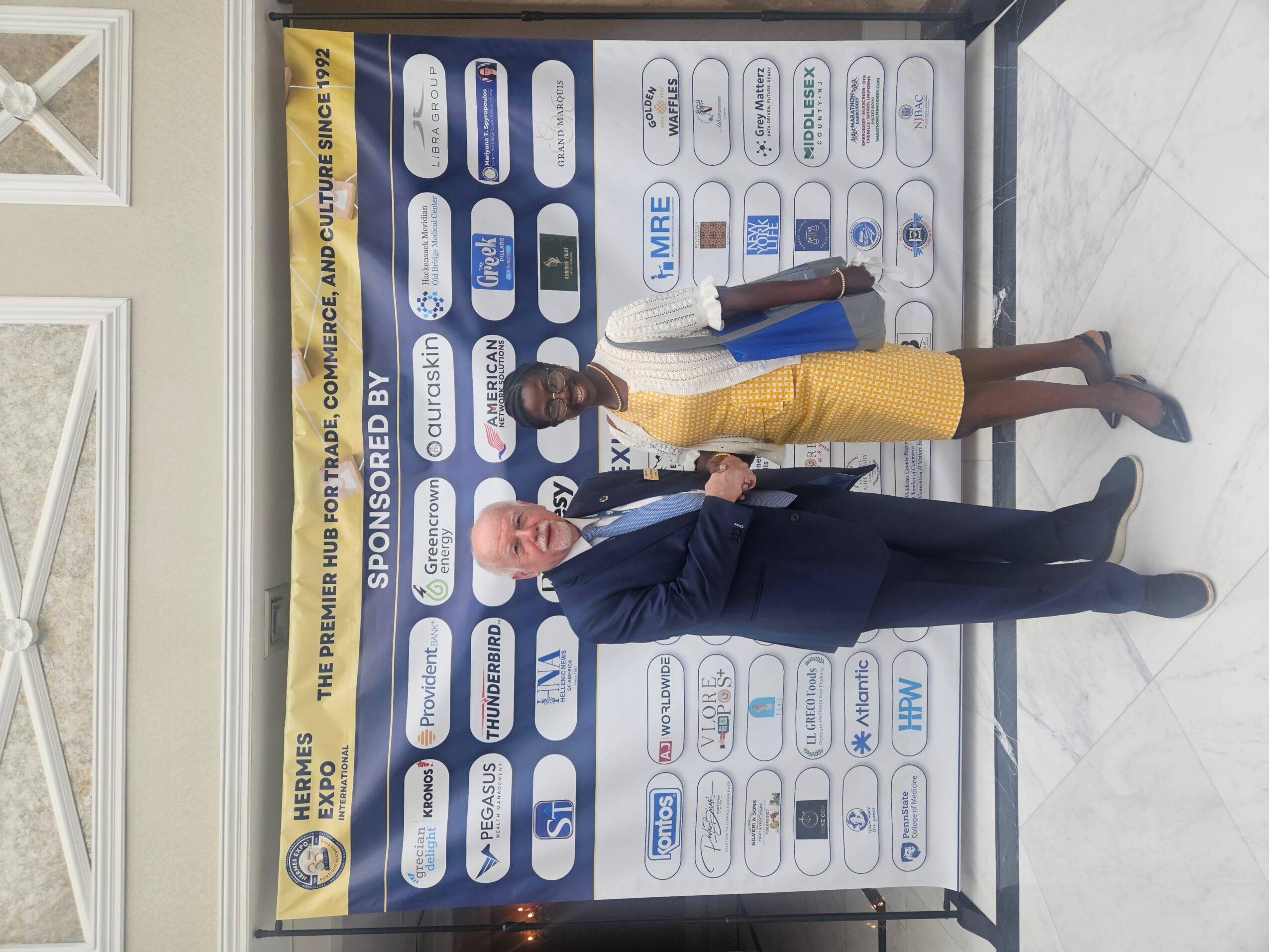 Two women standing in front of a sponsor banner, one in a yellow dress and the other in a dark suit, indoors.