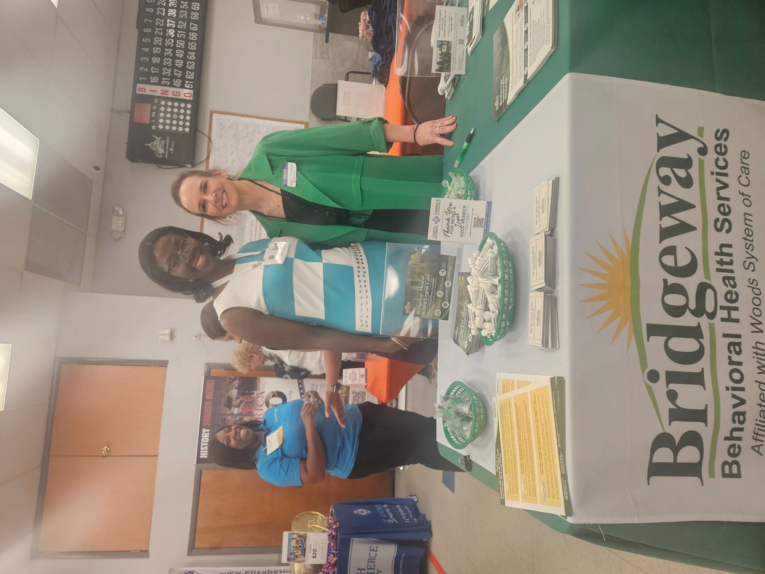 Two women stand behind a table with a cake and papers, at a Bridgeway Behavioral Health Services event, with a banner in the background.