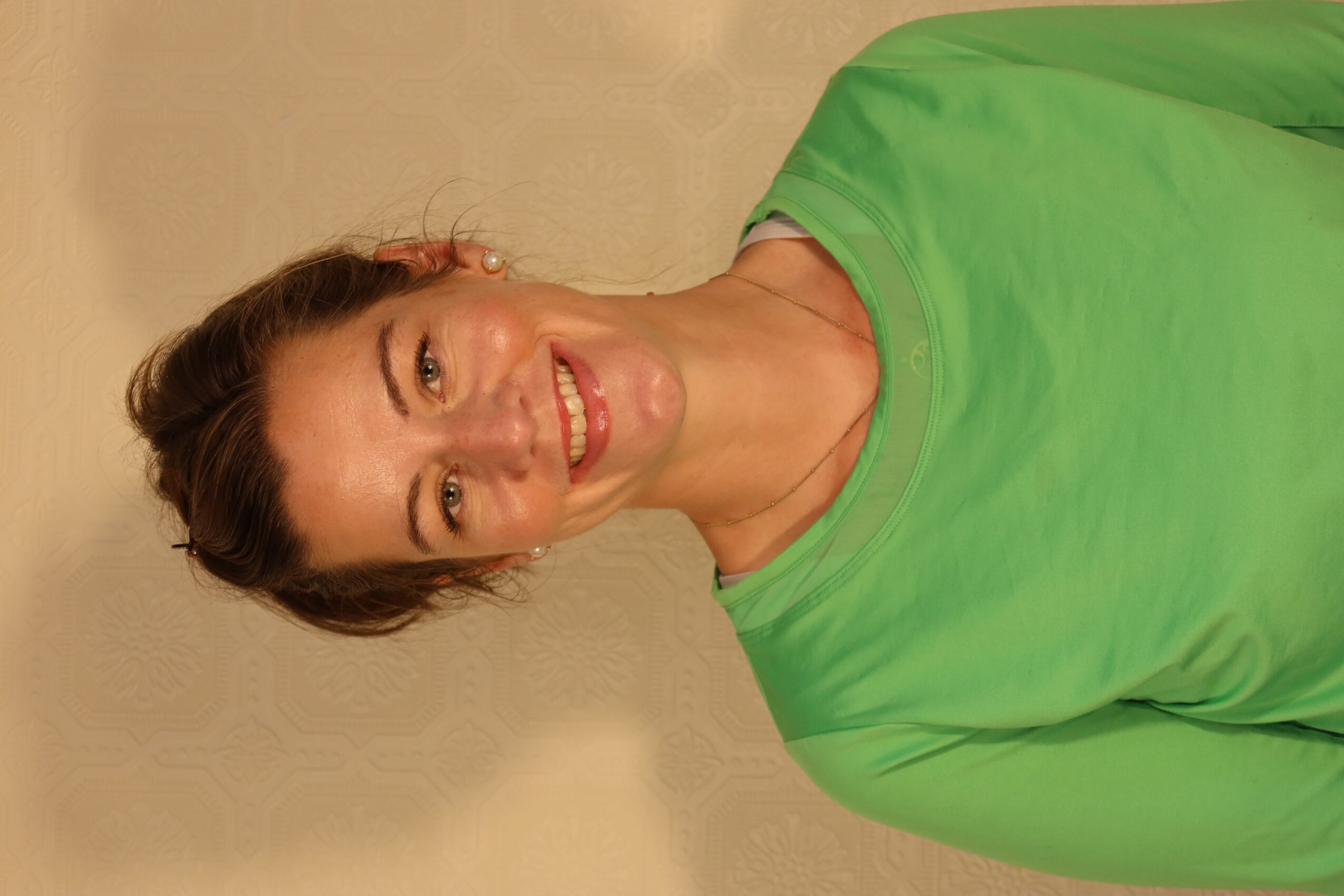 Woman with short brown hair smiling, wearing a green shirt, standing against a beige wall.