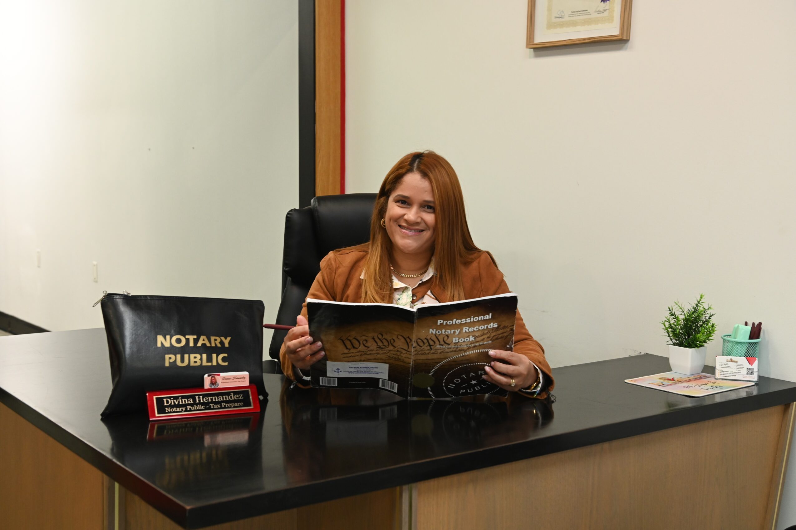 Woman with long hair sitting at desk, holding a brochure, smiling, with notary public sign on desk, office setting
