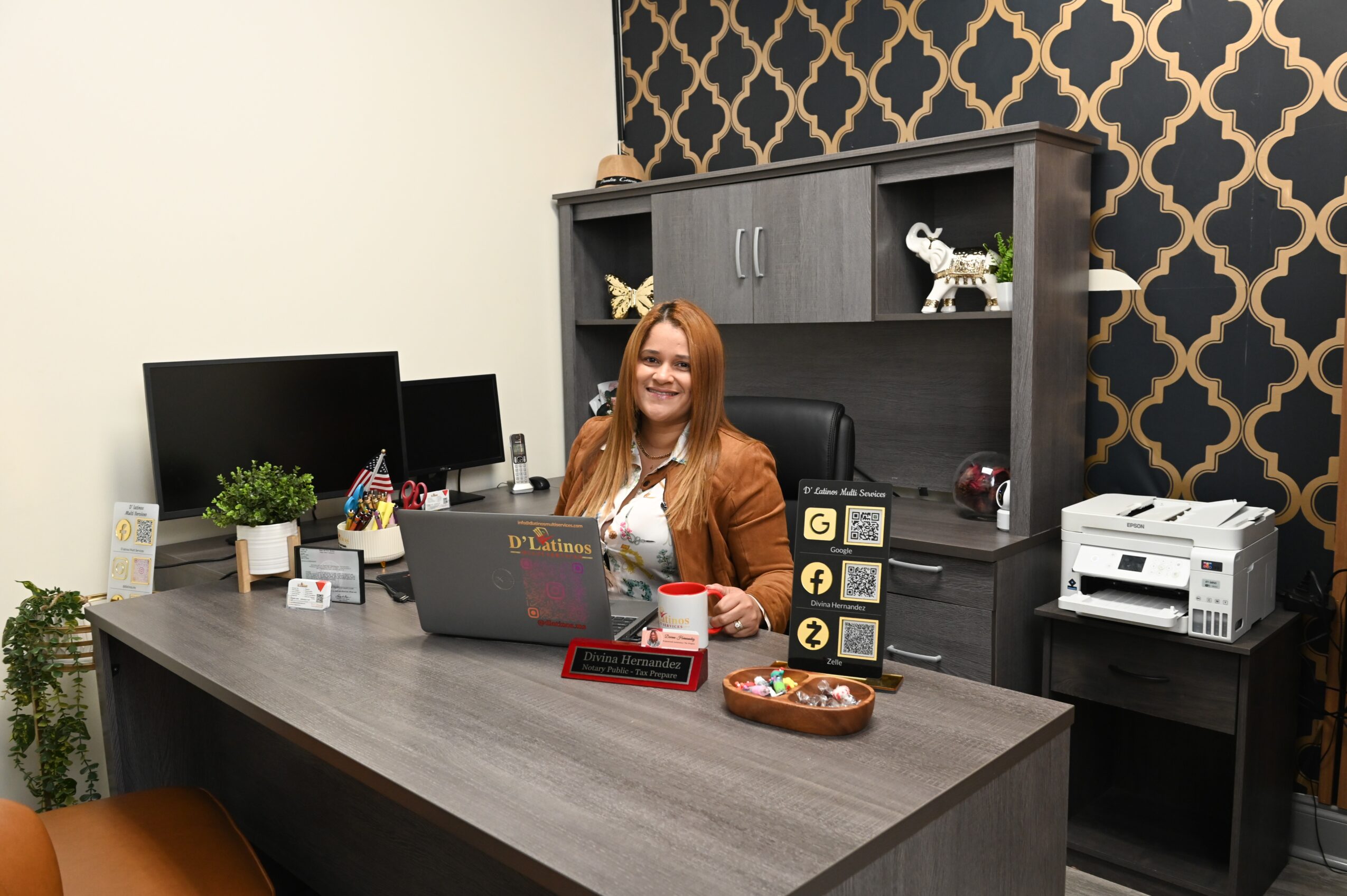 Woman sitting at desk with computer, bookshelf behind, patterned wall, and office supplies on desk.