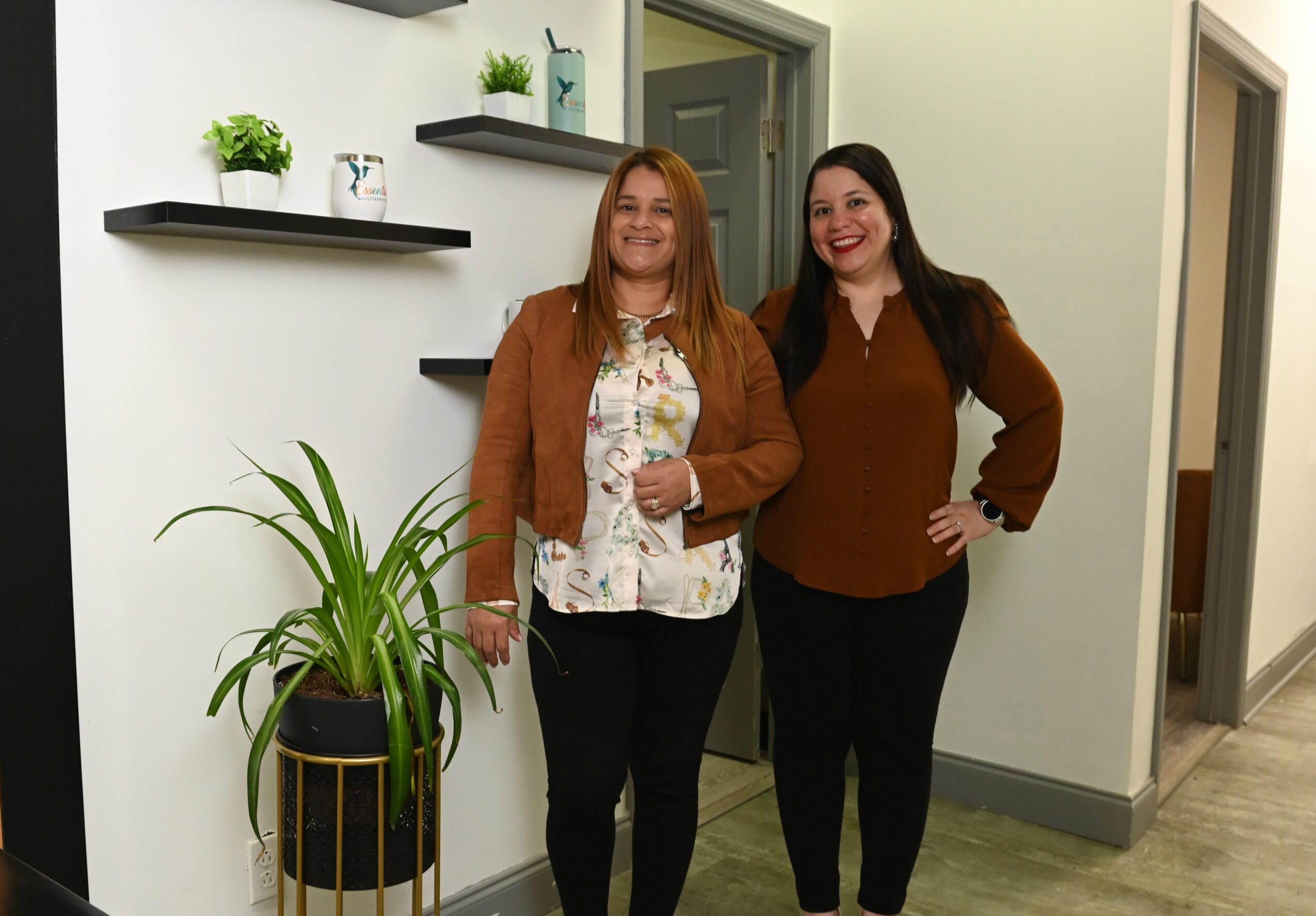 Two women standing indoors, smiling, with a plant and wall shelves behind them.
