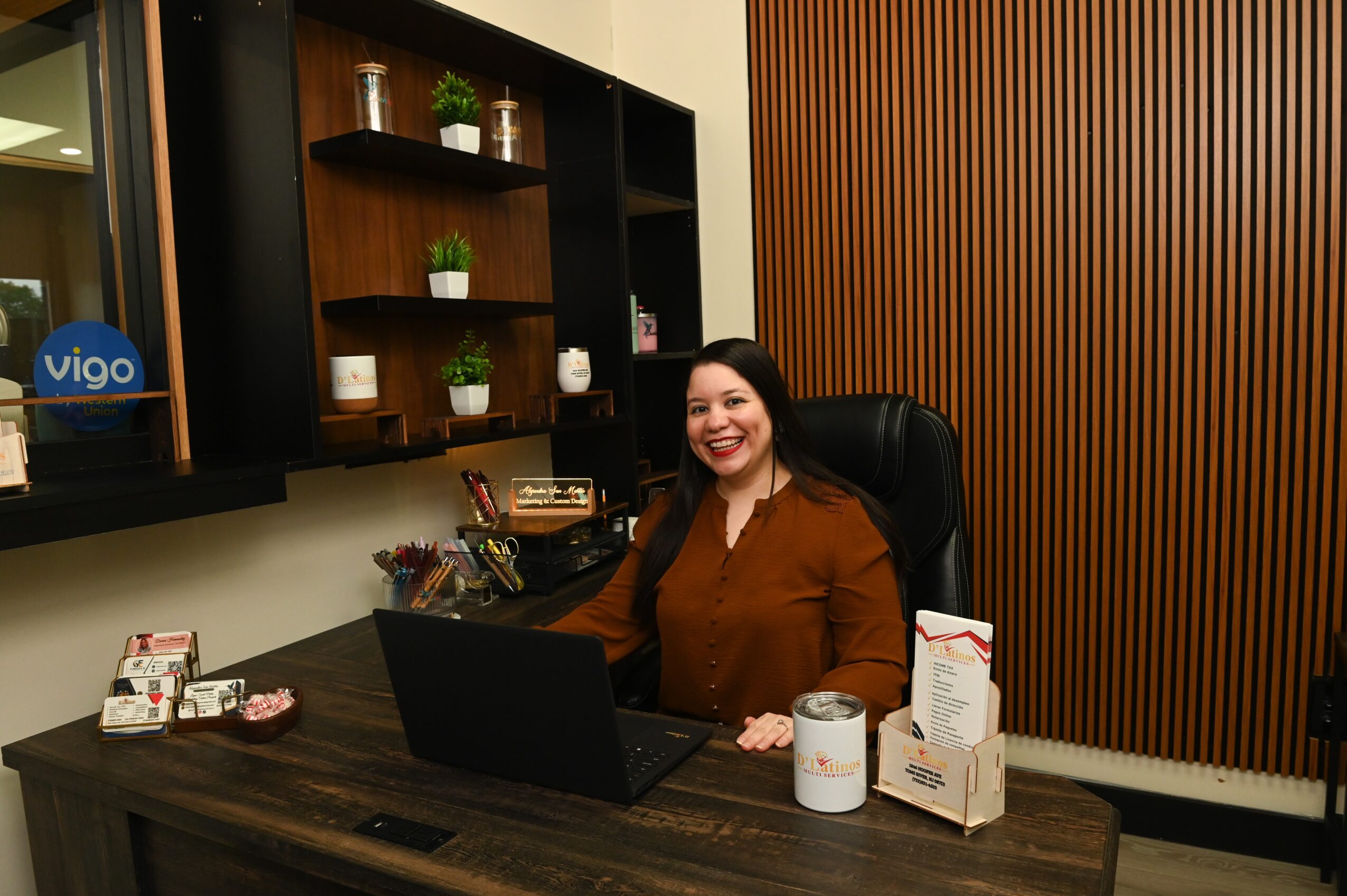 Woman sitting at a desk with a laptop, smiling, in an office with wooden wall panels and shelves.
