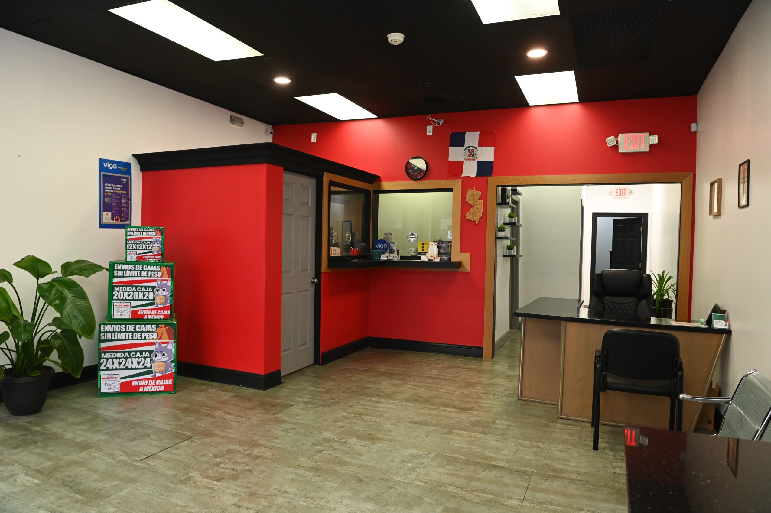 Reception area with red and white walls, a desk, a chair, and a magazine stand, with a plant in the corner.