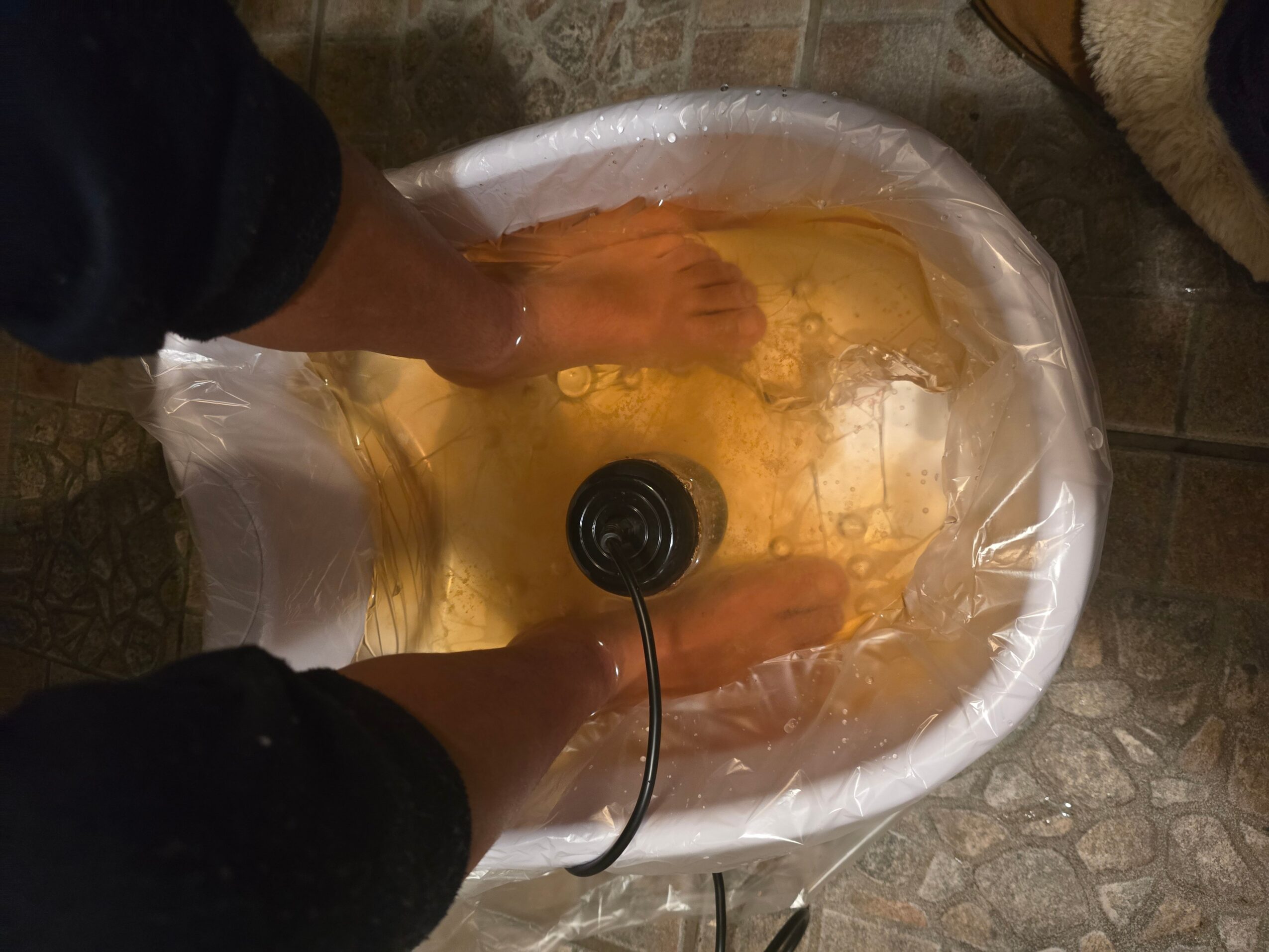 Person soaking feet in a plastic basin filled with water, with a black cord connected to a device in the basin.