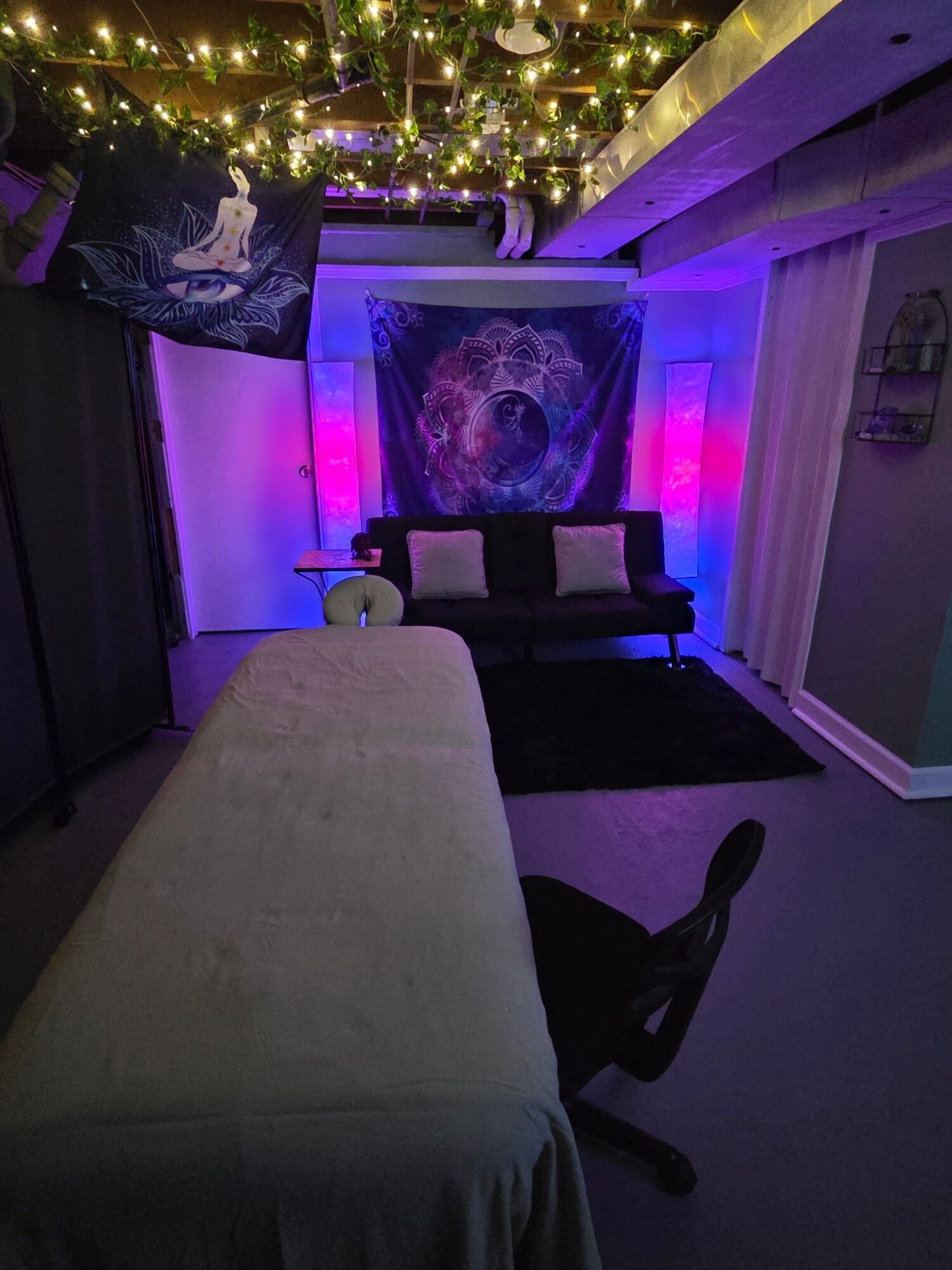 Room with a bed, chair, and wall art illuminated by purple and yellow lights, decorated with string lights and a pillow.