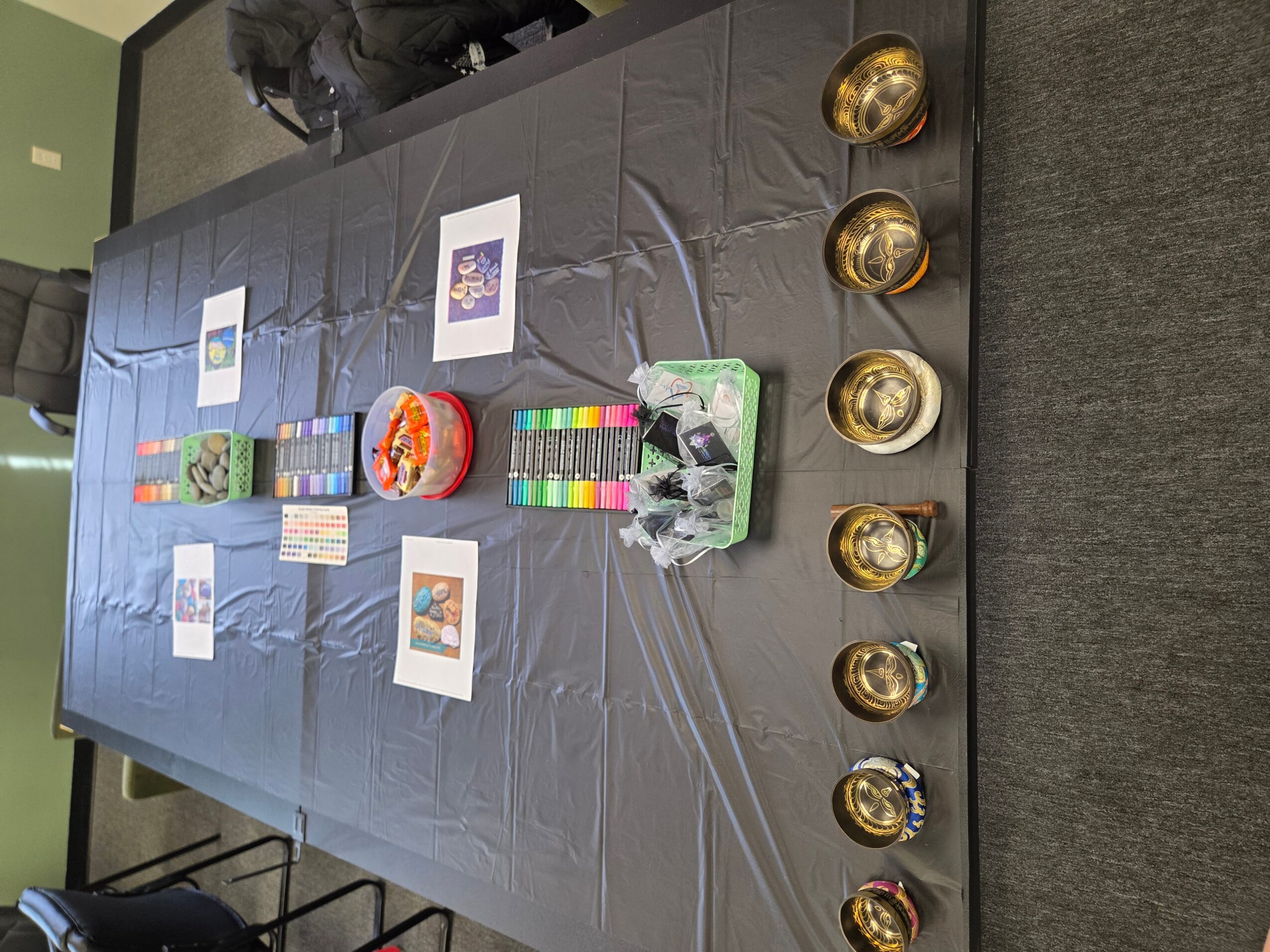 Table with colorful items, framed pictures, and small bowls, viewed from the side, with a gray carpeted floor.