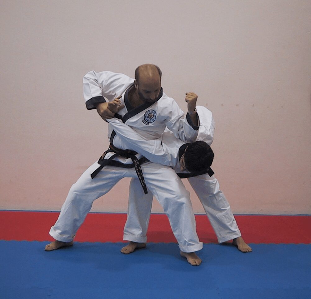 Two martial artists practicing a technique on a blue and red mat, one wearing a black belt and the other a white belt.