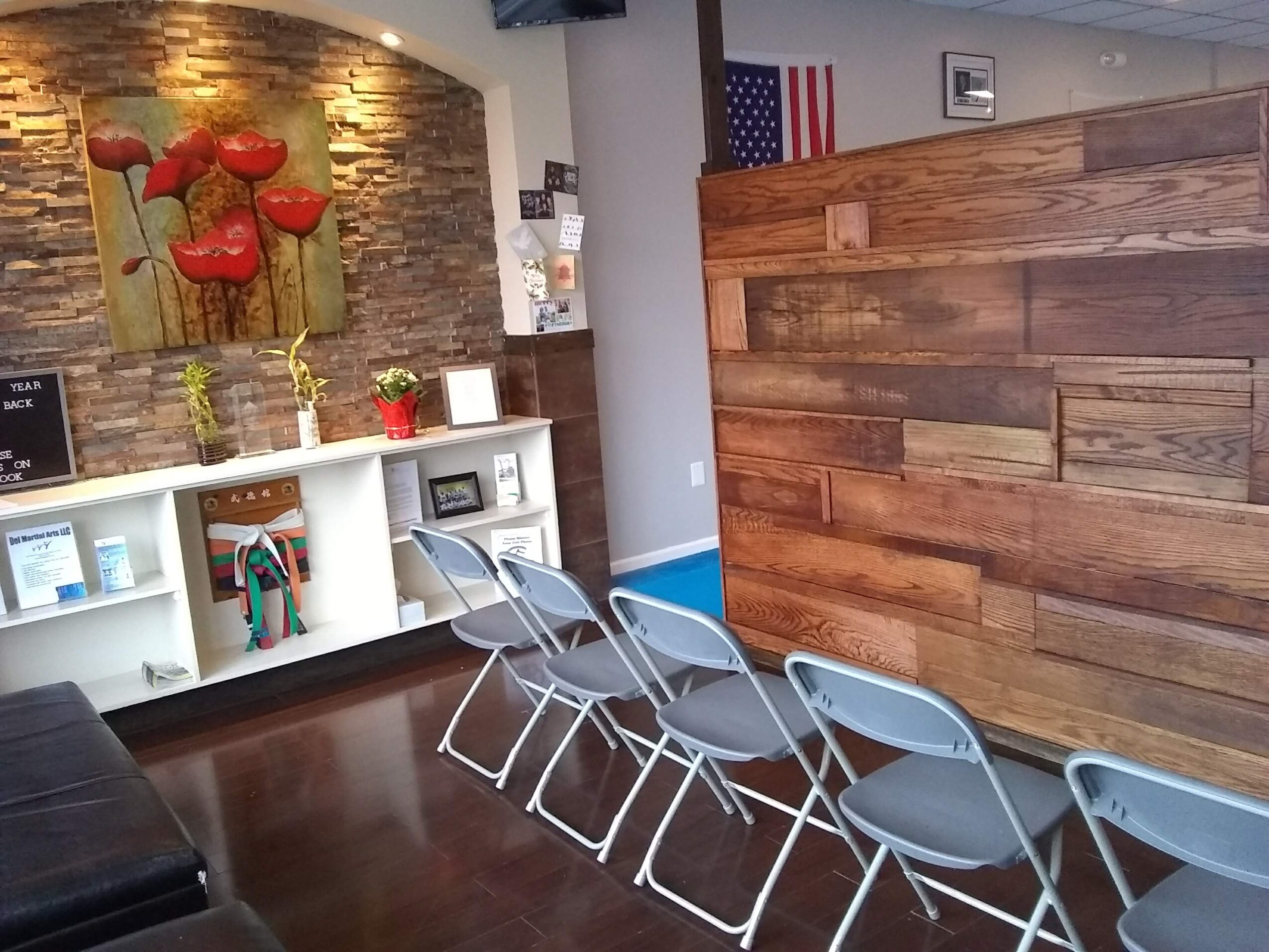 Waiting area with six gray chairs in front of a wooden wall, a white shelf with decorative items, and a stone wall with red flowers.
