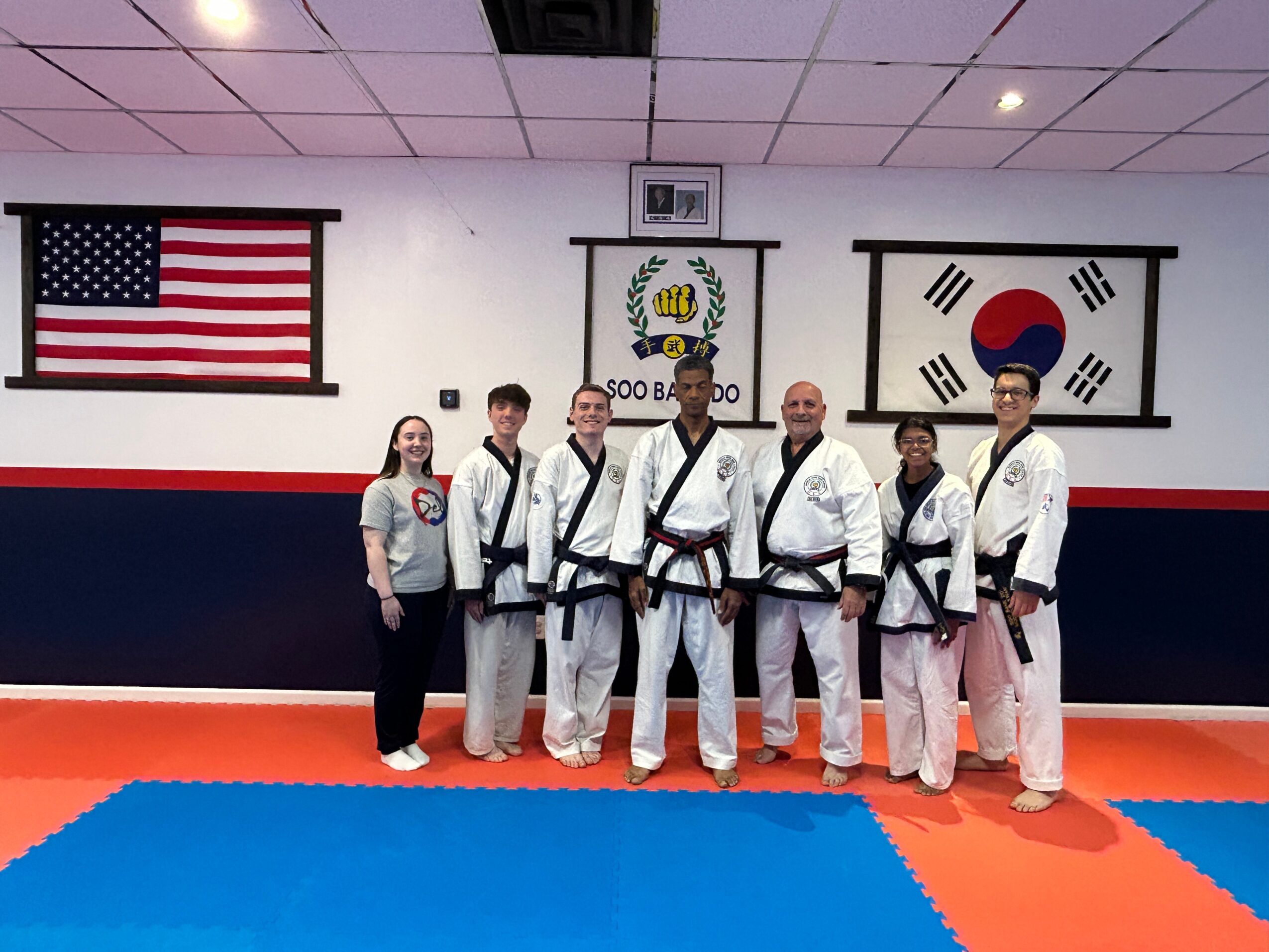 Group of six people in martial arts uniforms standing in a dojo with flags of the United States, Korea, and South Korea behind them.