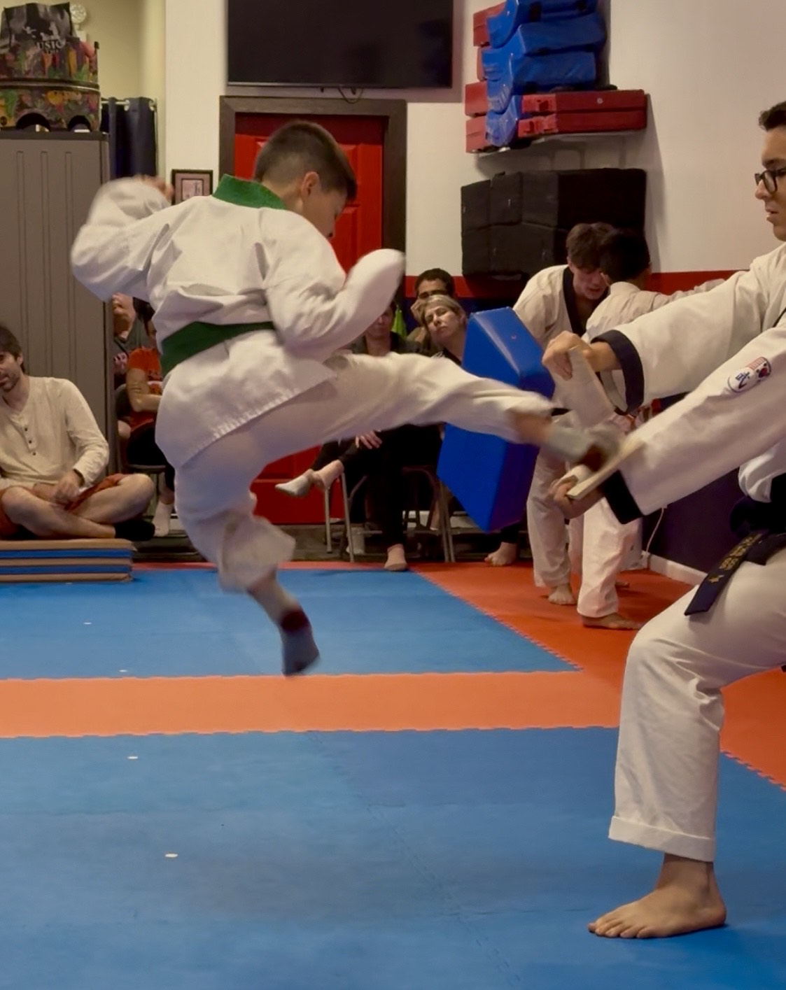 Child performing a high kick in martial arts uniform during a class, with spectators watching in the background.