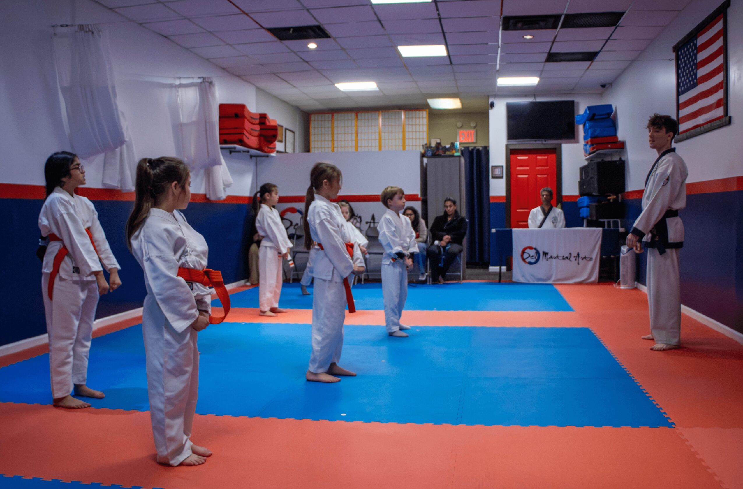 Children in martial arts uniforms standing in a line on a blue mat in a dojo, with an instructor nearby and an American flag on the wall.