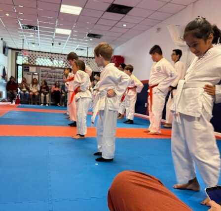 Children in martial arts uniforms standing in a line on a blue and orange mat, with spectators in the background.