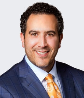 Man with dark curly hair, wearing a blue checkered suit and orange tie, smiling at the camera.