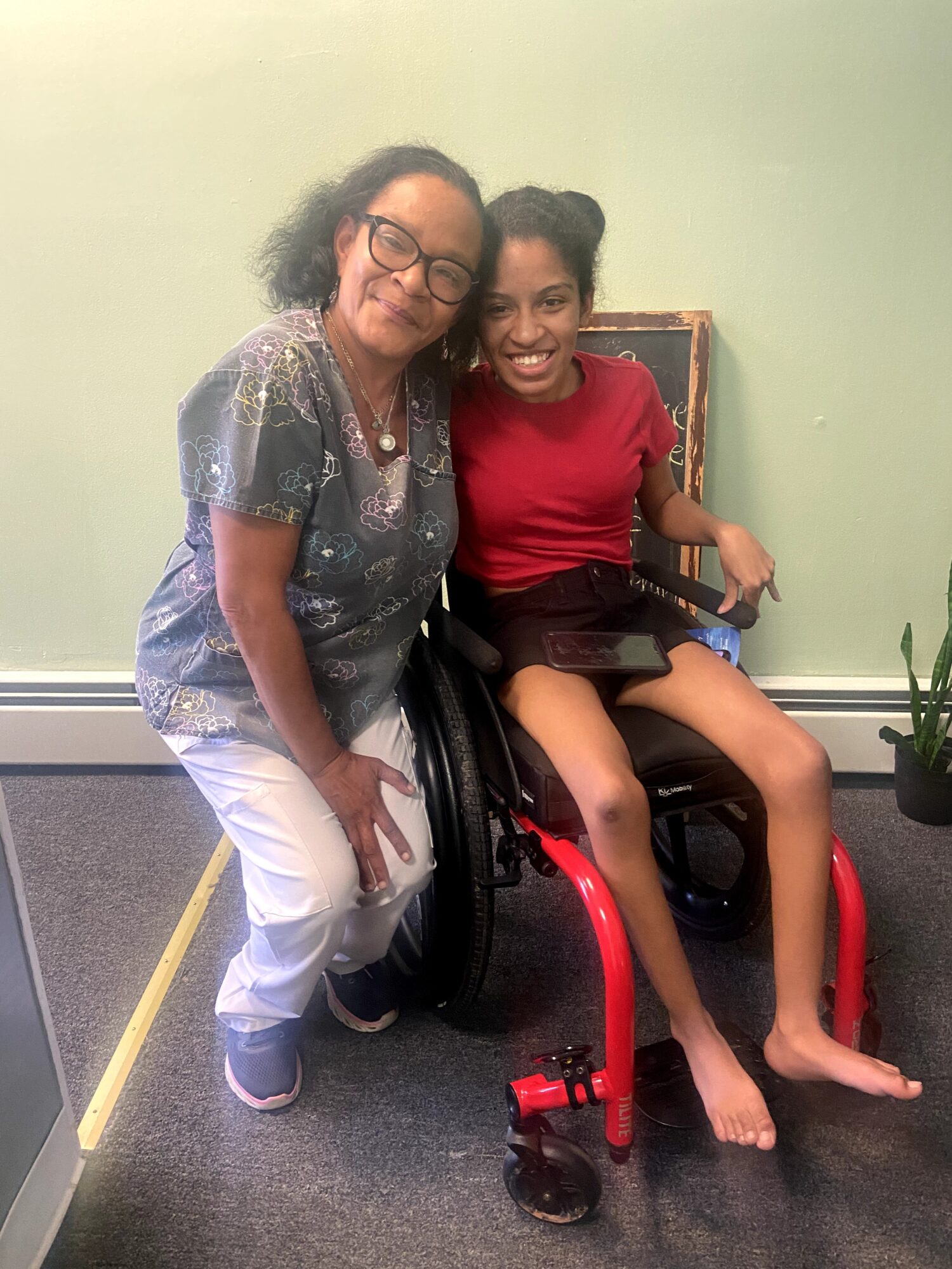 Two smiling females, one in a wheelchair, pose indoors against a light green wall, with a potted plant nearby.