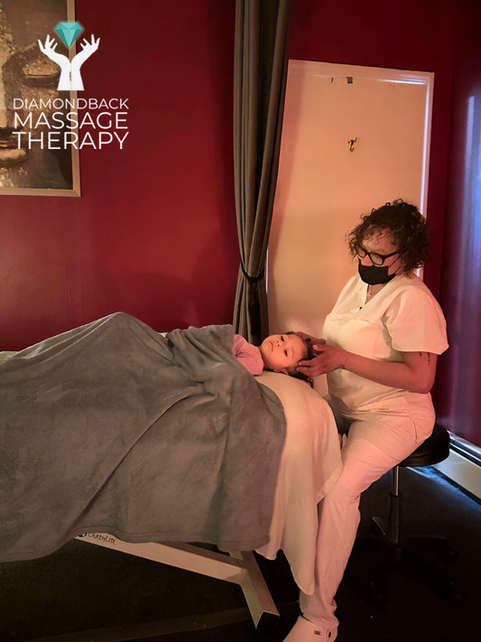 A massage therapist in white uniform and mask provides facial treatment to a person lying on a massage table, covered with a gray blanket, in a room with red walls and a window.