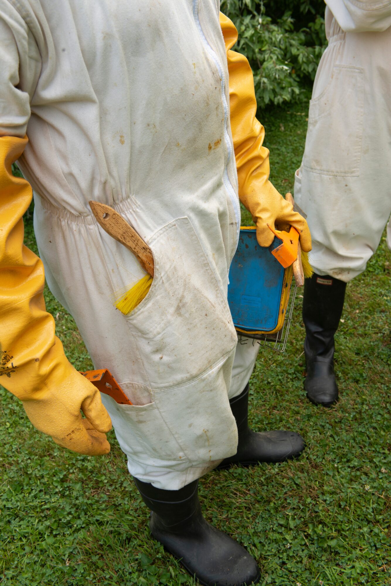 Two people in protective suits and gloves standing outdoors on grass, one holding a tool and a container.