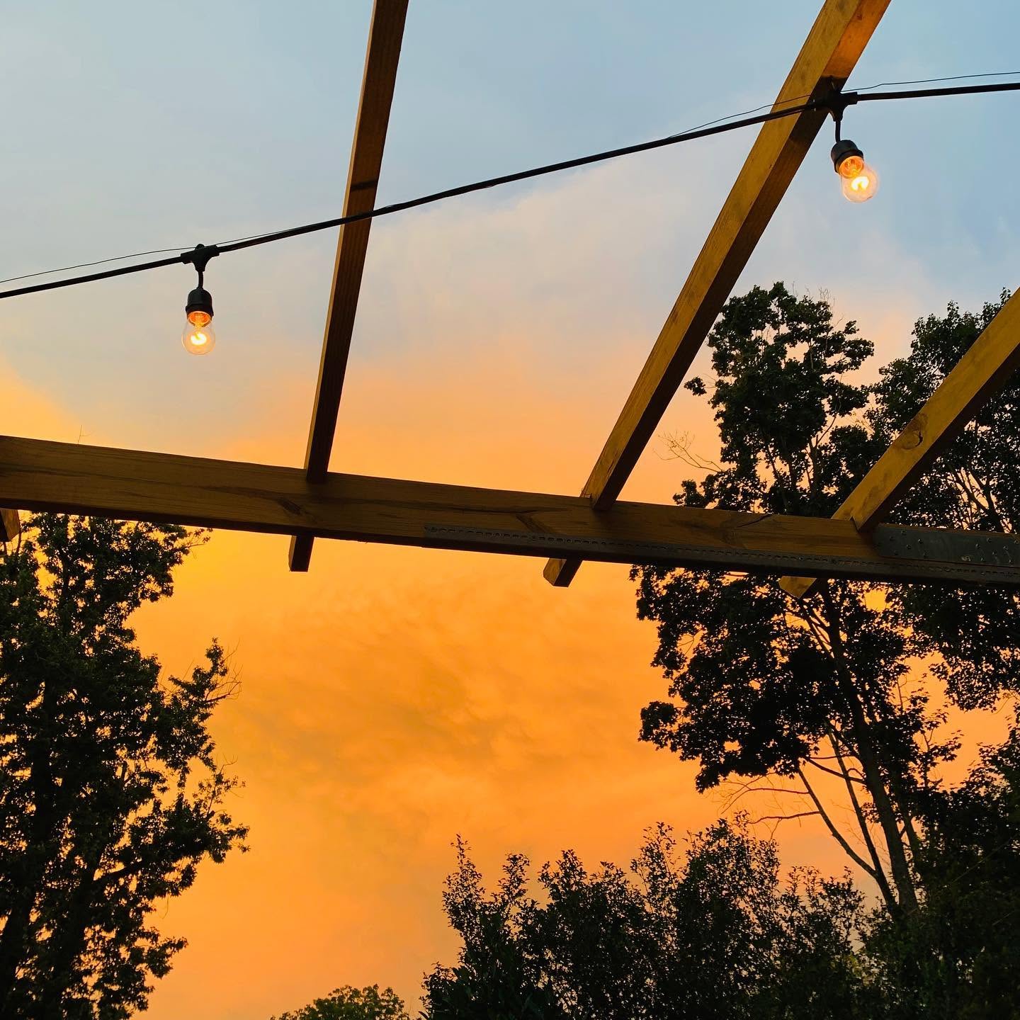 Sunset sky with orange and pink hues, silhouetted trees, and string lights hanging from a wooden structure.