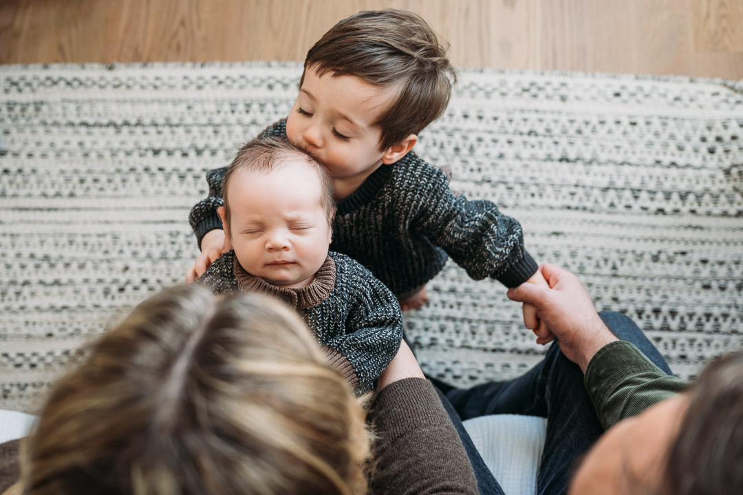 Two children, one older and one younger, sitting on a patterned rug indoors, with the older child holding the younger.