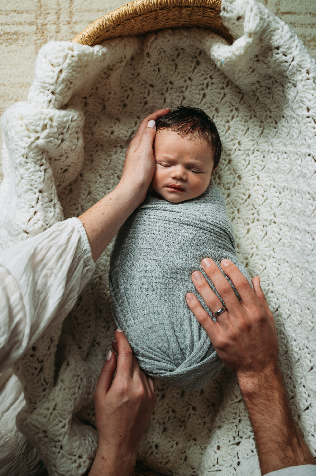 Baby wrapped in a blanket lying on a blanket, with two adults gently touching the baby's head and body.