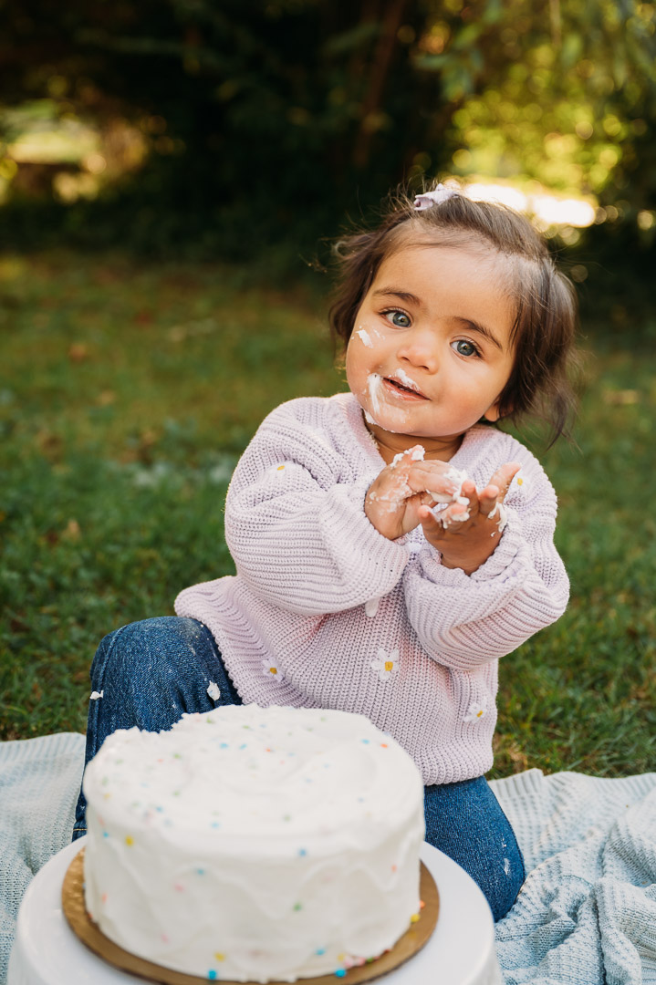 Child with dark hair and a light sweater, sitting outdoors with a cake, smiling and holding cake in hands.
