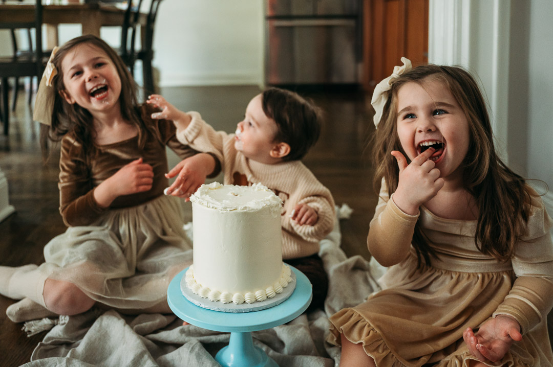 Three children sitting on the floor celebrating with a cake, smiling and laughing, in a cozy room.