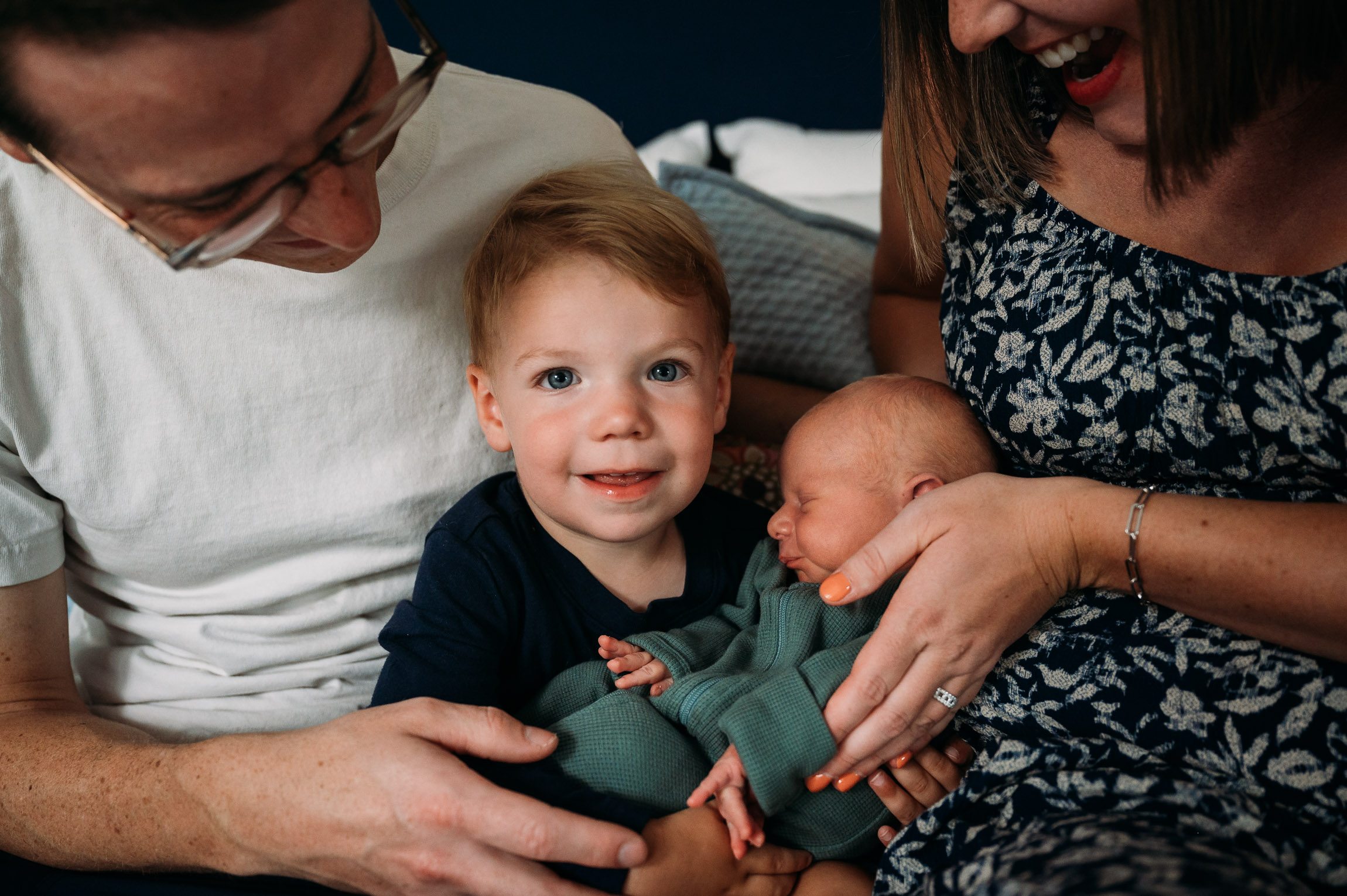 Family with a young child and newborn, smiling and holding the babies, indoors. Word count: 20.