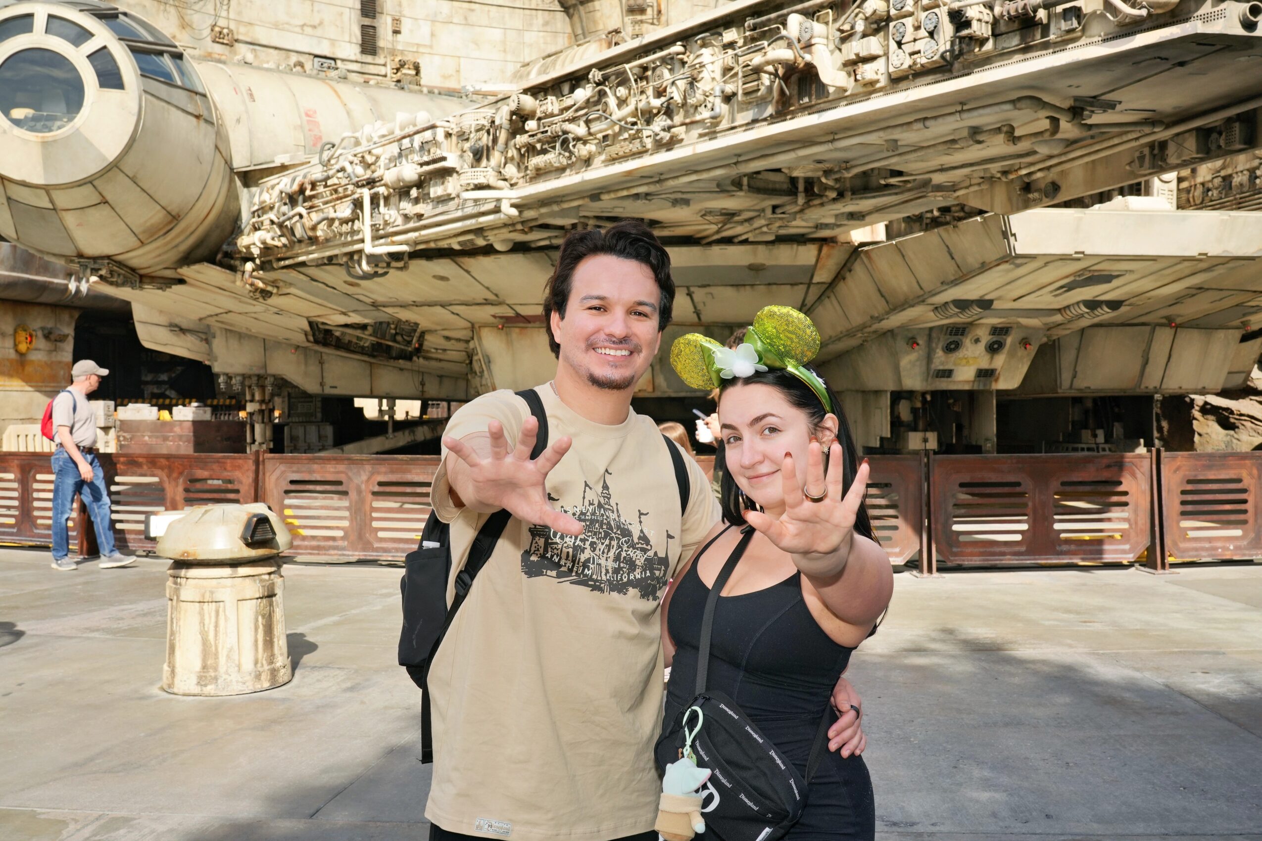 Two people smiling and posing in front of a spaceship, one holding a ball, under a large spacecraft structure.