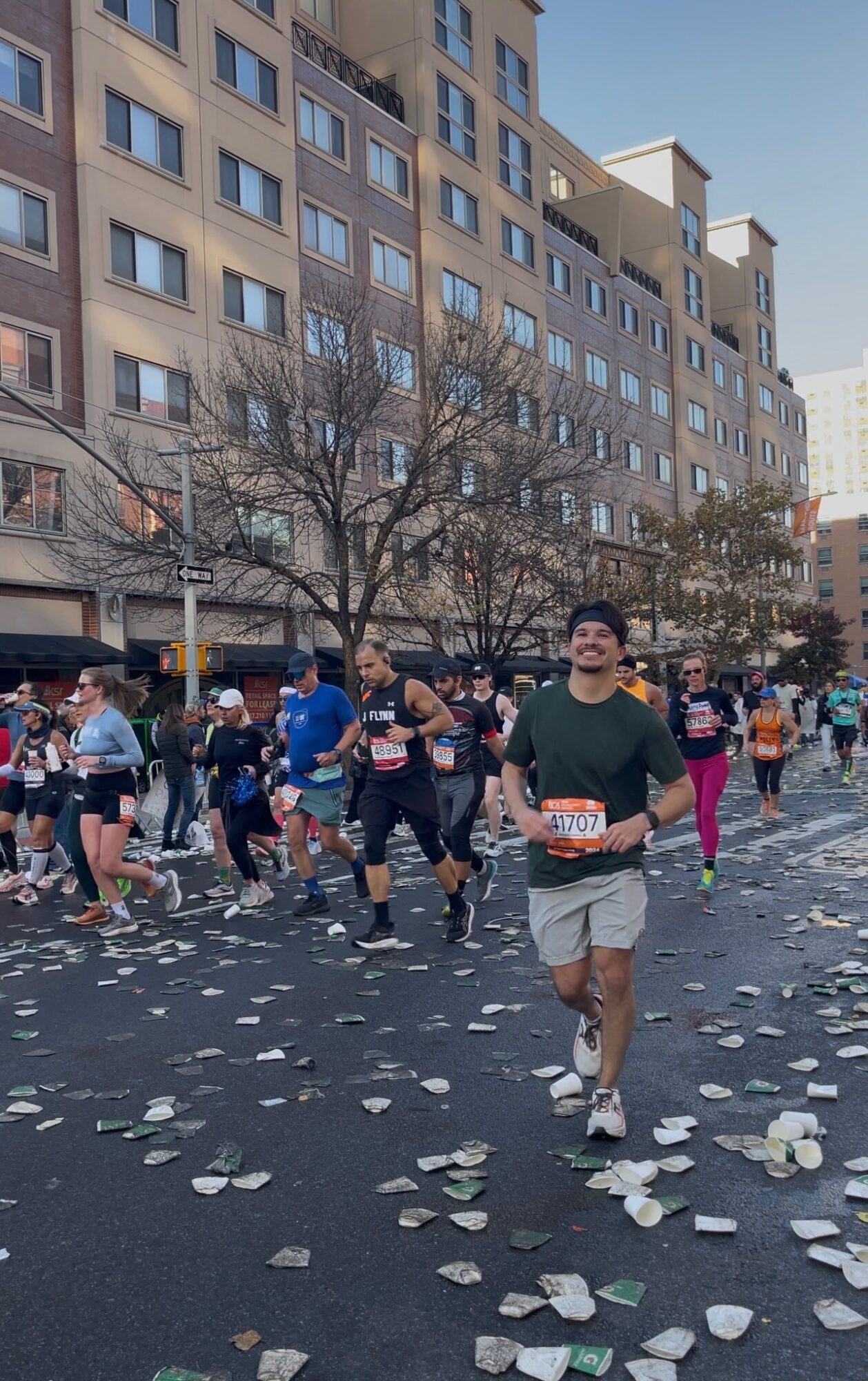 Runner in green shirt and shorts participating in a race on city street with other runners and tall buildings in background.