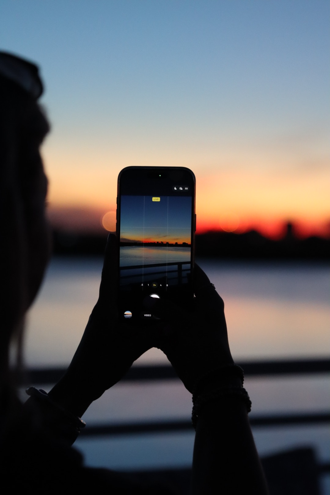 Person taking a photo of a sunset with a smartphone, silhouette visible, colorful sky, water in background.