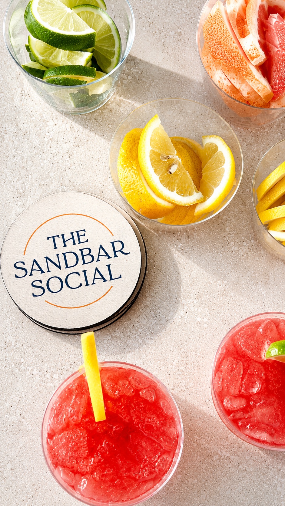 Lemon slices in a bowl, watermelon slices, and a container labeled 'The Sandbar Social' on a light surface.