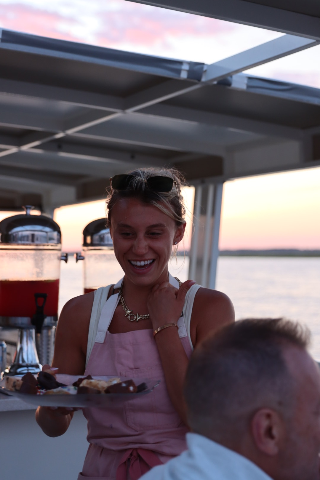 Smiling woman with sunglasses on her head, wearing a pink top, sitting outdoors with a sunset in the background.