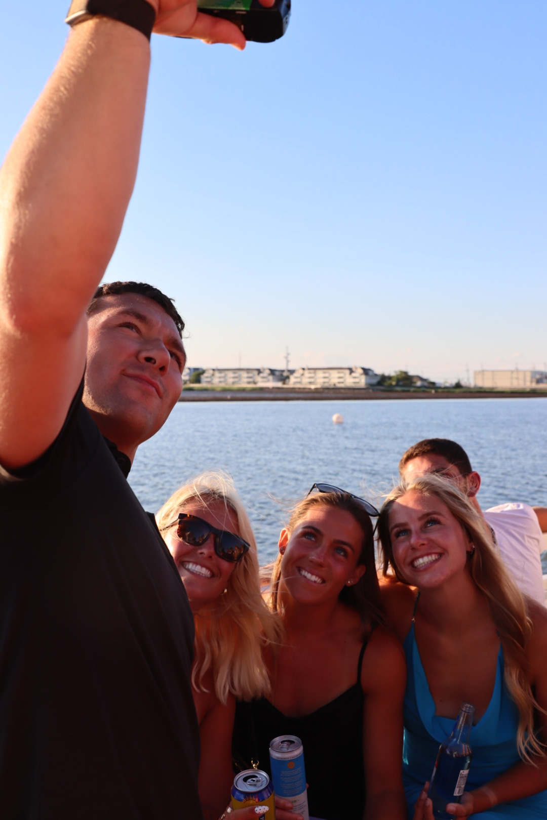 Group of four people smiling and taking a selfie near a body of water with a cityscape in the background.