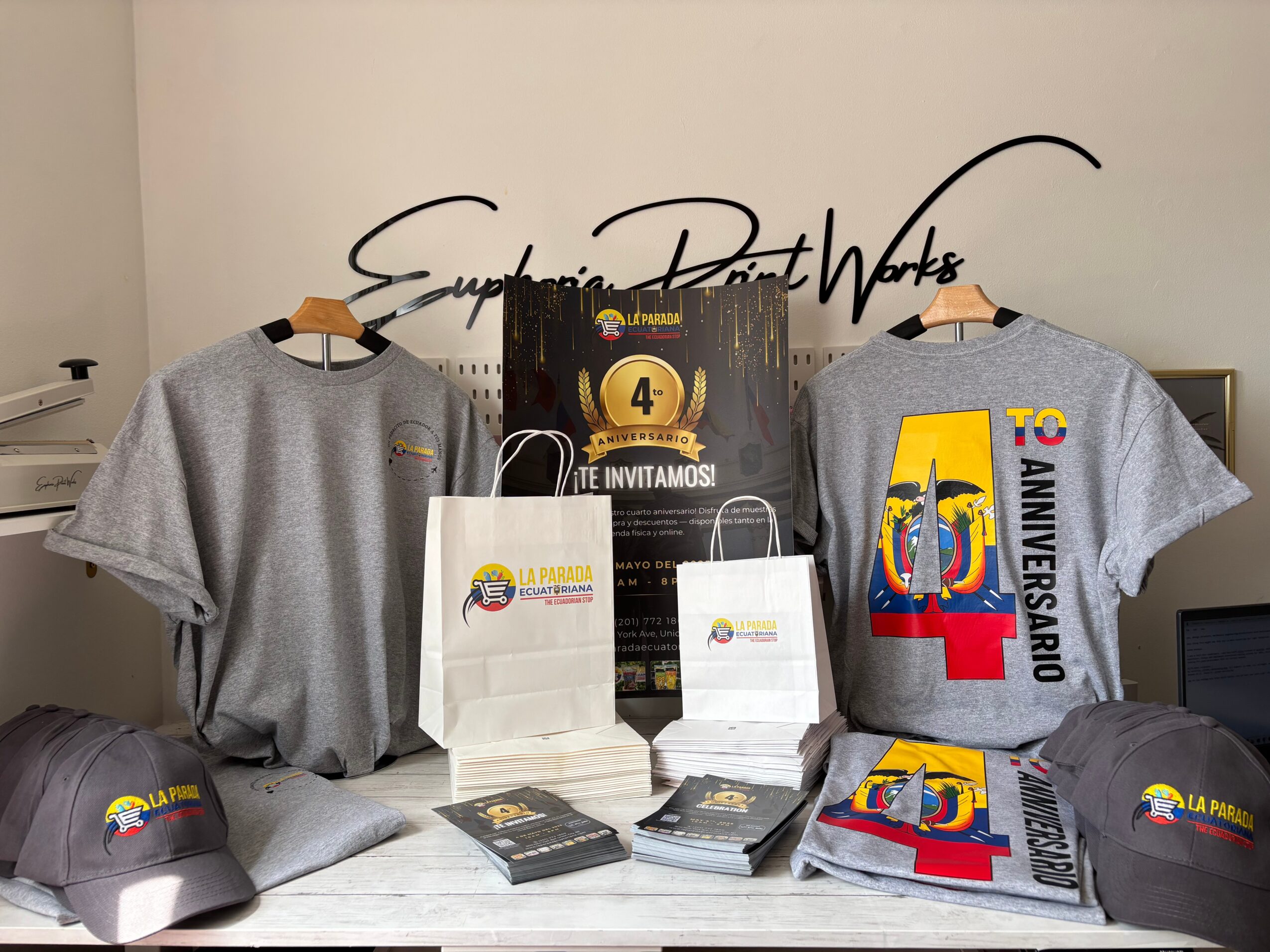 Two gray T-shirts with colorful designs, hats, and gift bags on a table, with a wall and sign in the background.